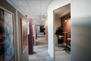 Couloir de bureau moderne avec cabines de travail modulaires et art encadré. Espace détente à droite : mur texturé, table haute, tabourets en liège et éclairage ciblé.