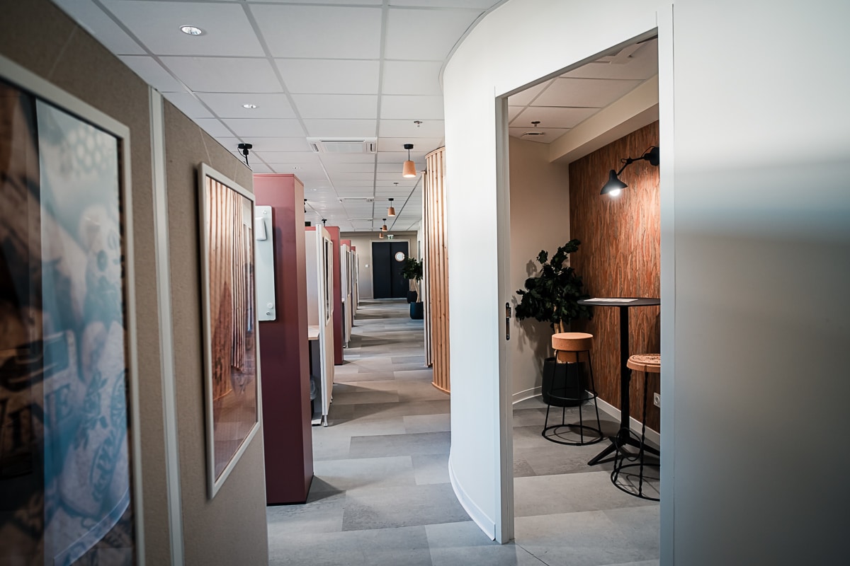 Couloir de bureau moderne avec cabines de travail modulaires et art encadré. Espace détente à droite : mur texturé, table haute, tabourets en liège et éclairage ciblé.