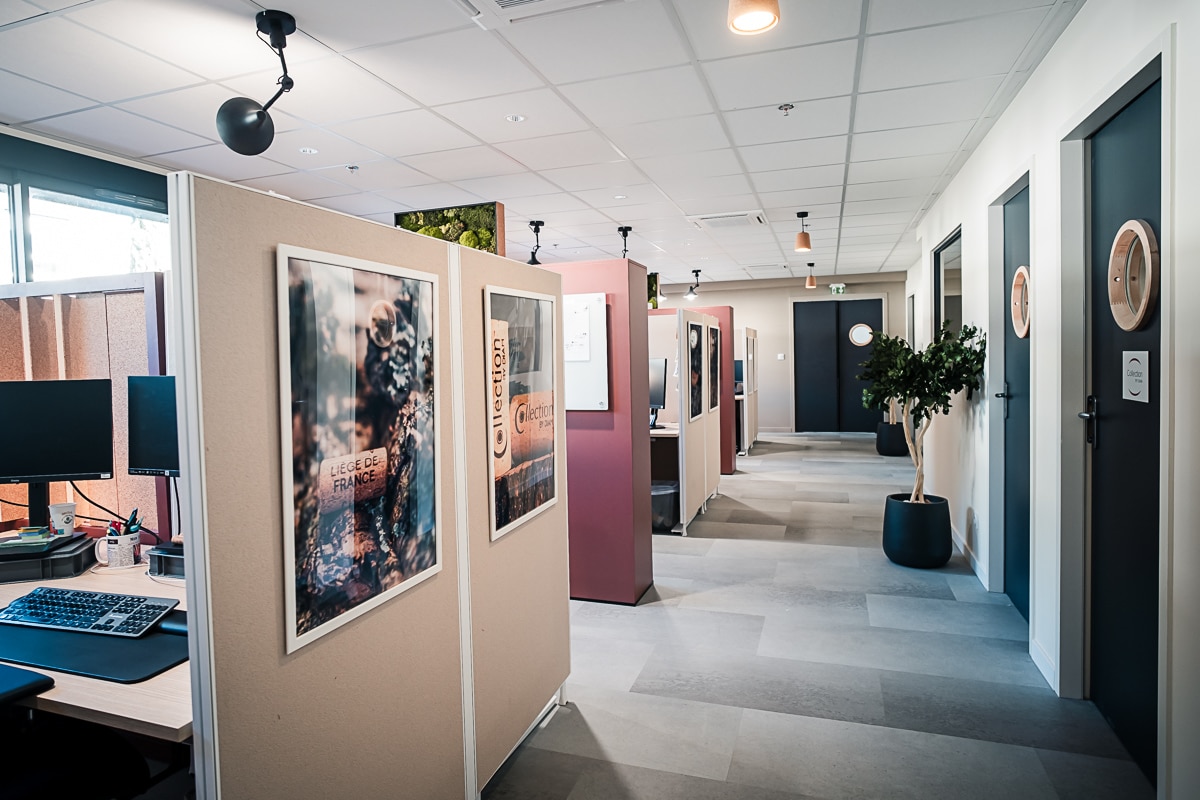 Couloir de bureau moderne avec cloisons modulaires et postes de travail. Design épuré, portes sombres à hublots et affiches "LIEGE DE FRANCE".