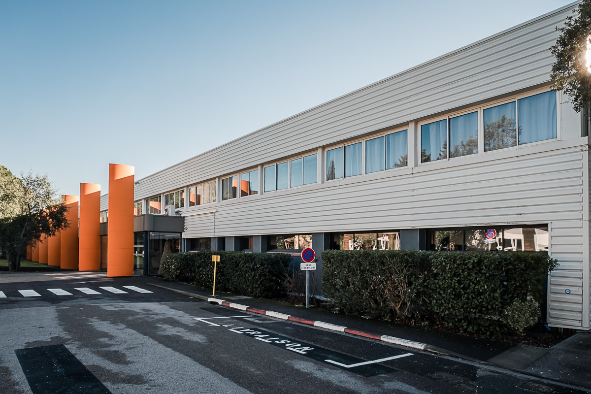 Façade de bureaux moderne à bardage blanc, avec une série de hauts piliers orange vif marquant l'entrée, jouxtant une zone de livraison et un panneau indiquant : "SAUF LIVRAISONS".