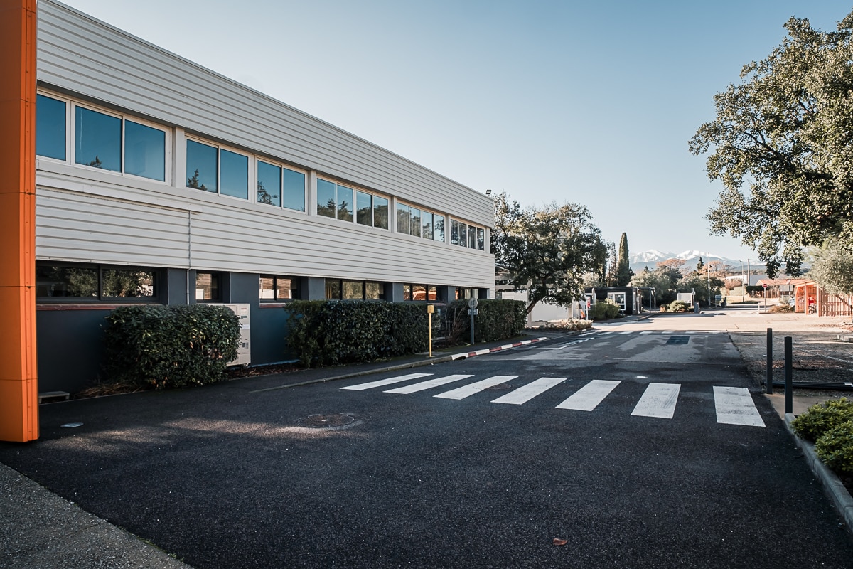 Bâtiment de bureaux moderne, bordé de haies. Passage piéton vers l'entrée sécurisée, avec vue dégagée sur les montagnes enneigées.