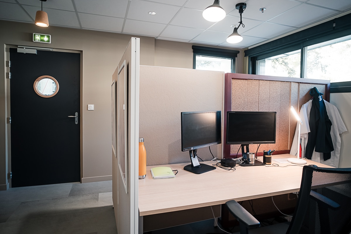 Espace de travail moderne. Deux moniteurs sur un bureau clair près d'une porte noire avec hublot et une signalétique d'évacuation verte.