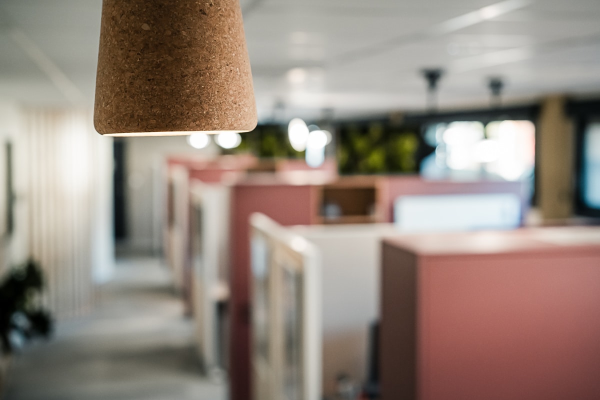 Lampe suspendue en liège éclairant un open space moderne. Rangs de bureaux aux cloisons roses et blanches, lumière douce.