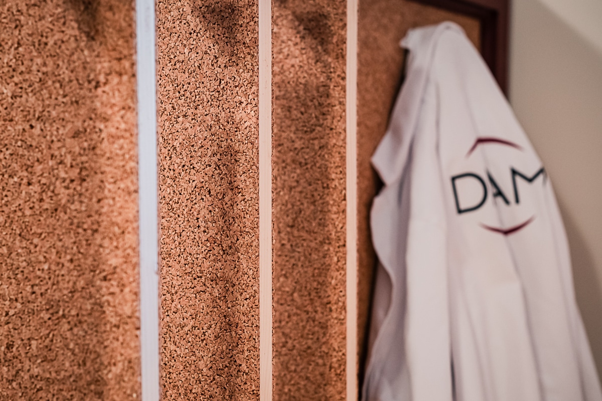 Panneaux muraux en liège granulé formant des bandes verticales. À droite, une blouse de laboratoire blanche est accrochée, affichant le logo 'DAM'.