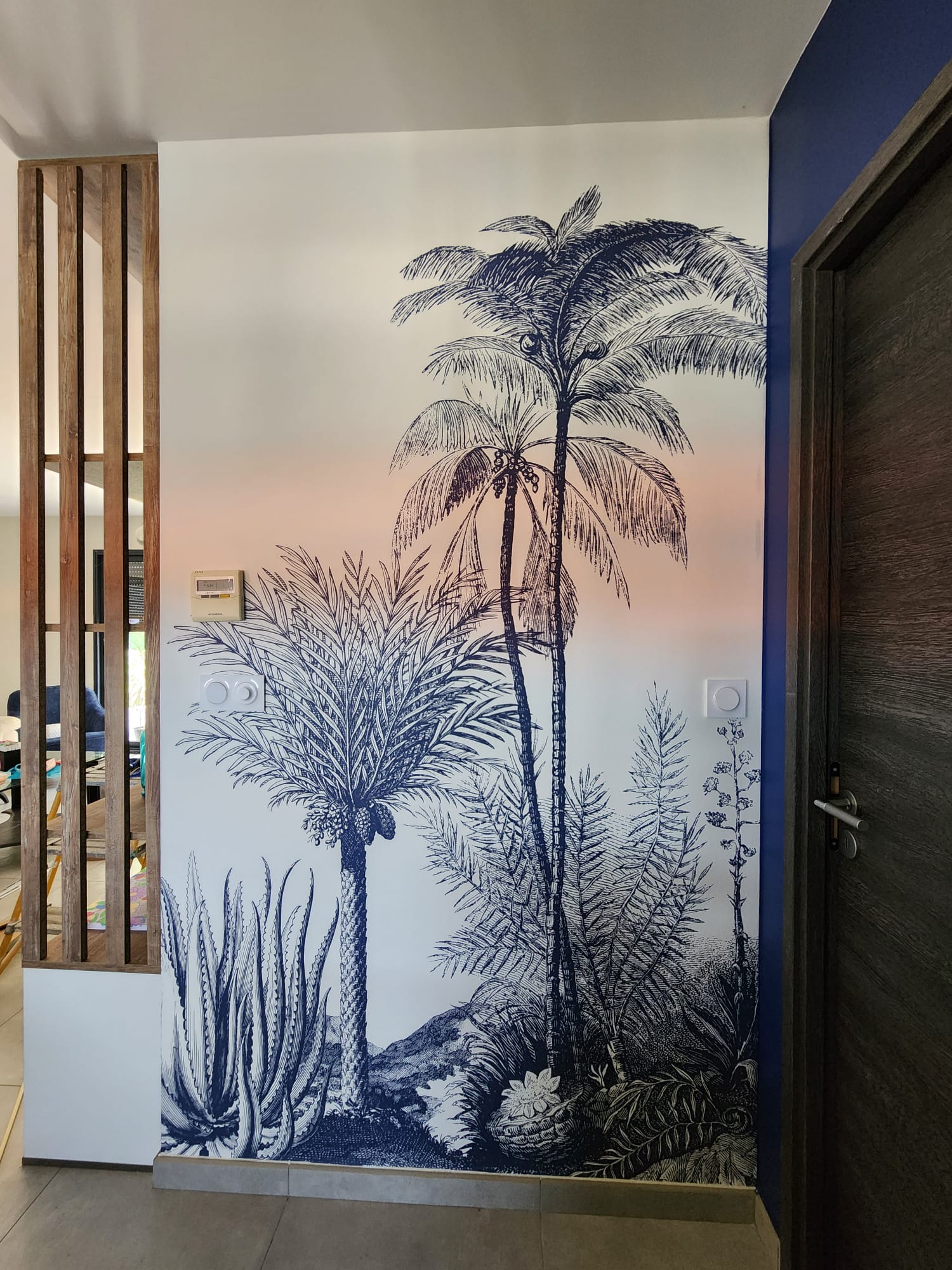 Papier peint bleu motif jungle avec palmiers et agave sur mur blanc entre bois et porte.