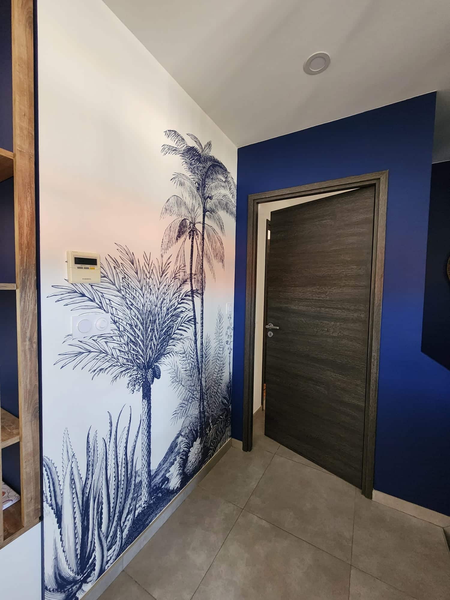 Papier peint tropical bleu avec palmiers sur mur blanc à côté d'une porte sombre et d'un mur bleu nuit.