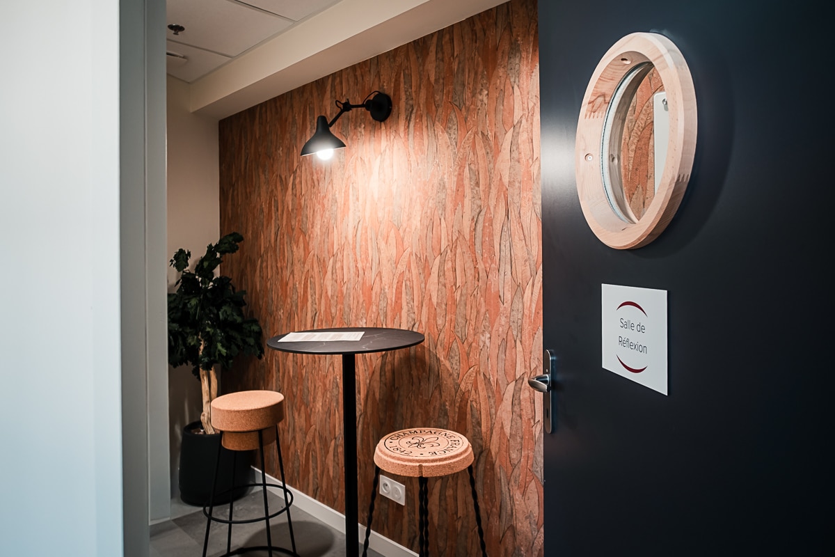 Nook avec mur texturé terre cuite. Table haute, tabourets en liège gravés Champagne. Lampe design. Porte noire : "Salle de Réflexion".
