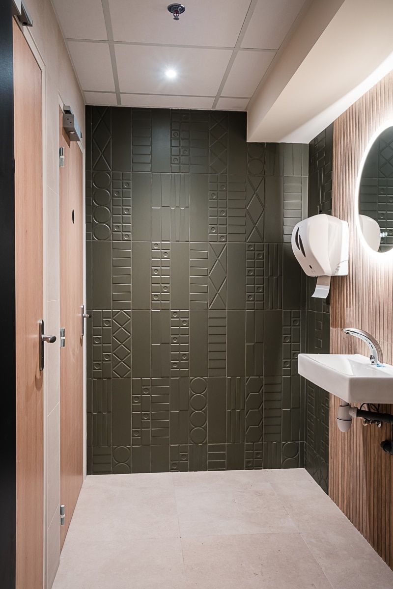 Design de toilettes avec mur vert en carrelage texturé, lavabo blanc, distributeur, miroir lumineux ovale et portes de cabines en bois clair.
