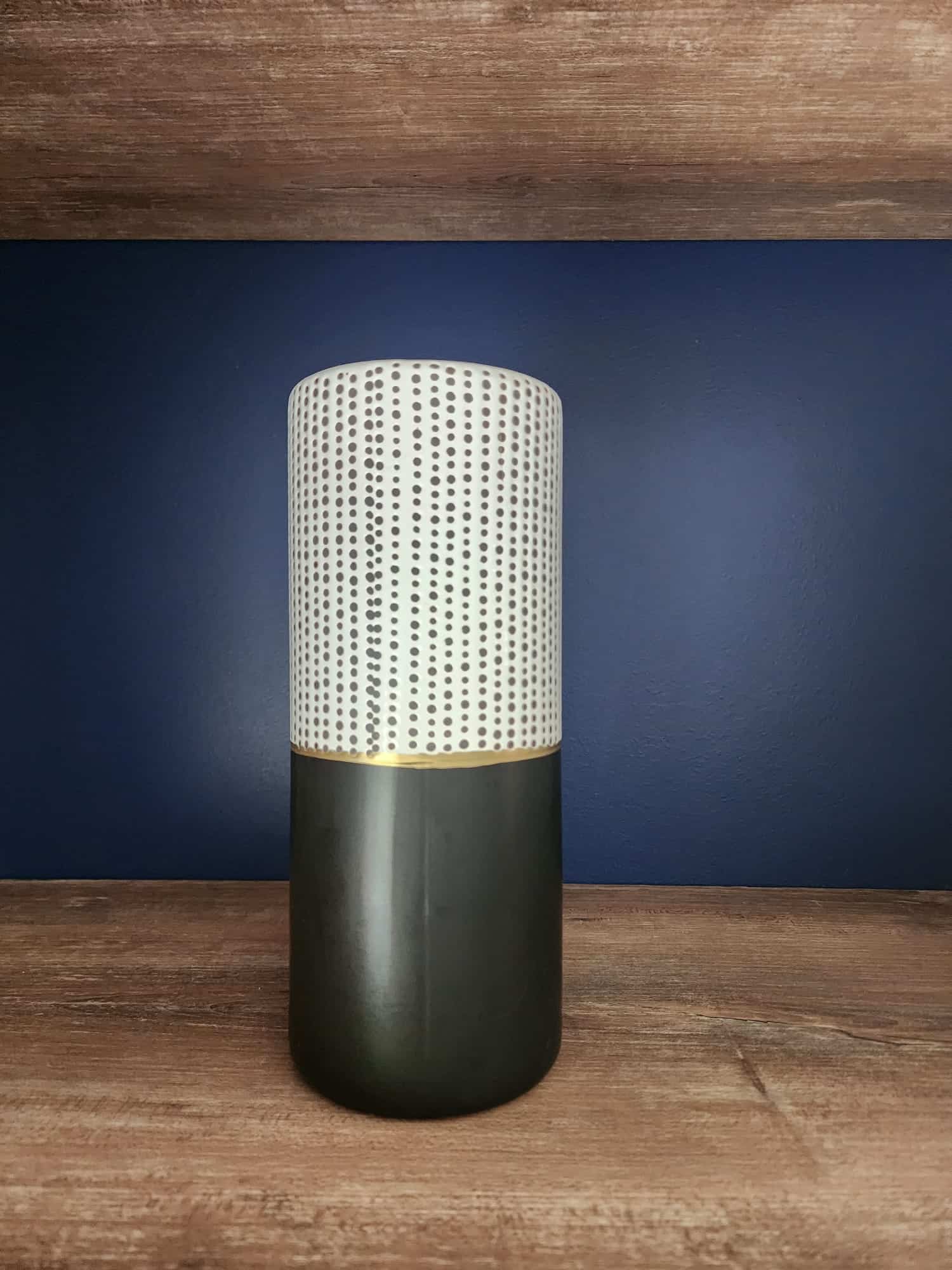Vase bicolore blanc à pois et noir mat avec liseré doré sur étagère en bois et fond bleu foncé.