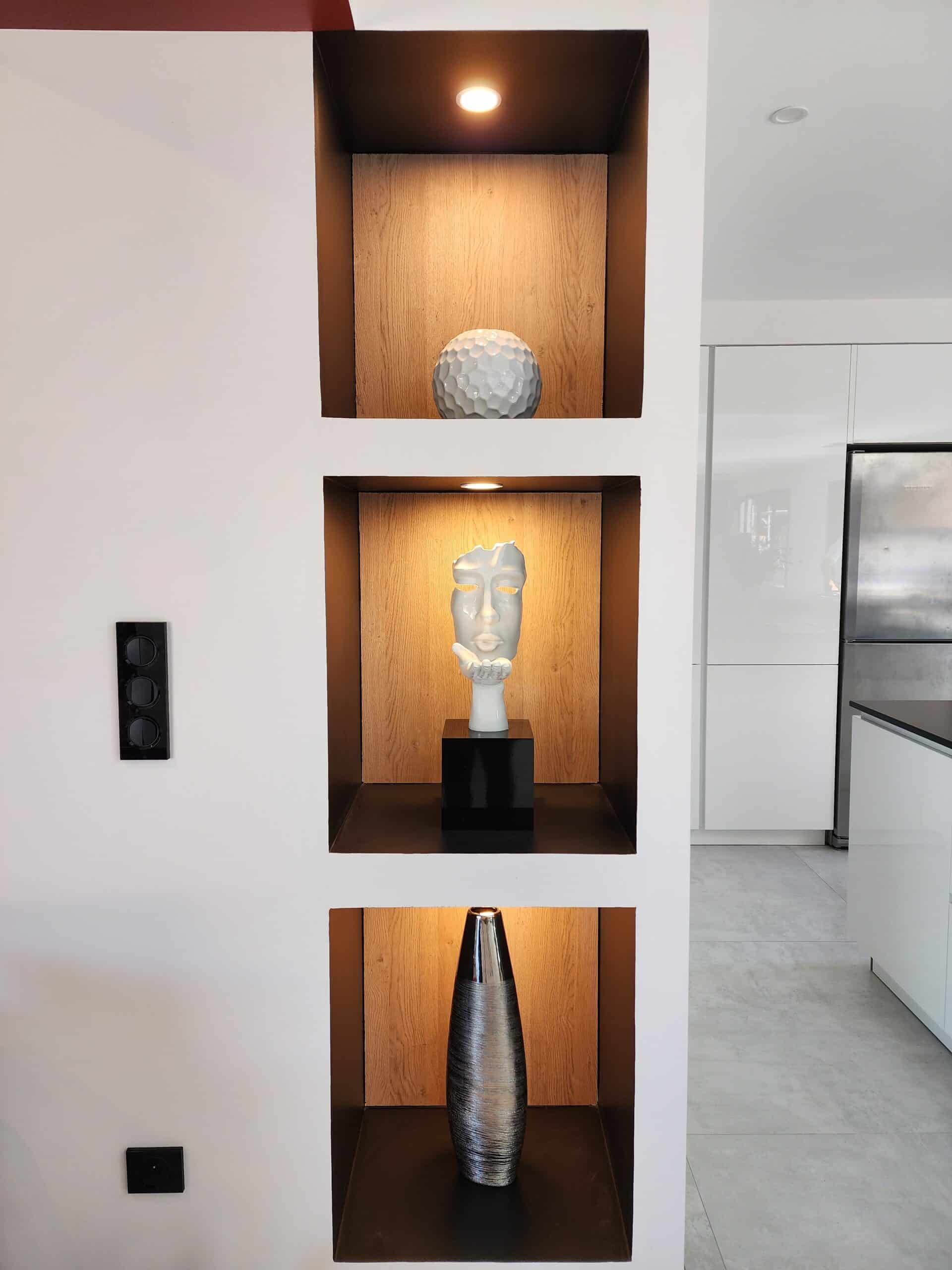 Niches murales éclairées avec sculpture de visage, vase argenté et boule déco dans un intérieur moderne.