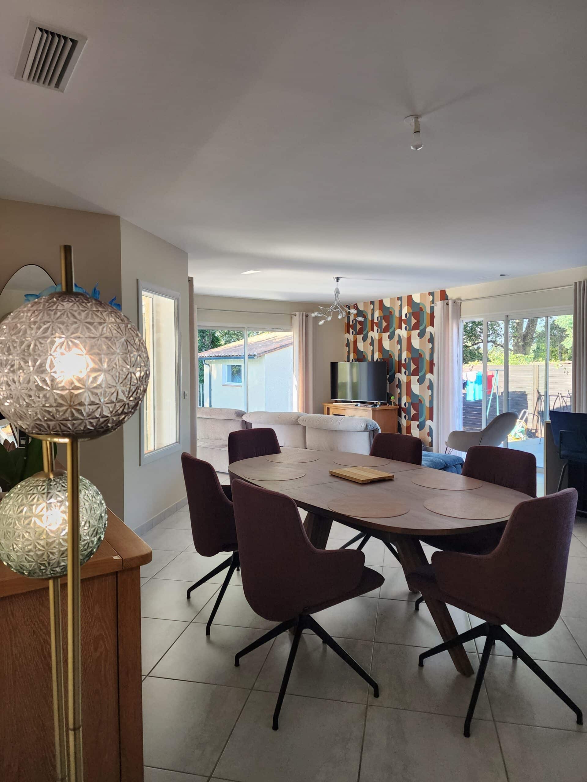 Salle à manger moderne, table ovale, chaises bordeaux et salon avec papier peint géométrique coloré.