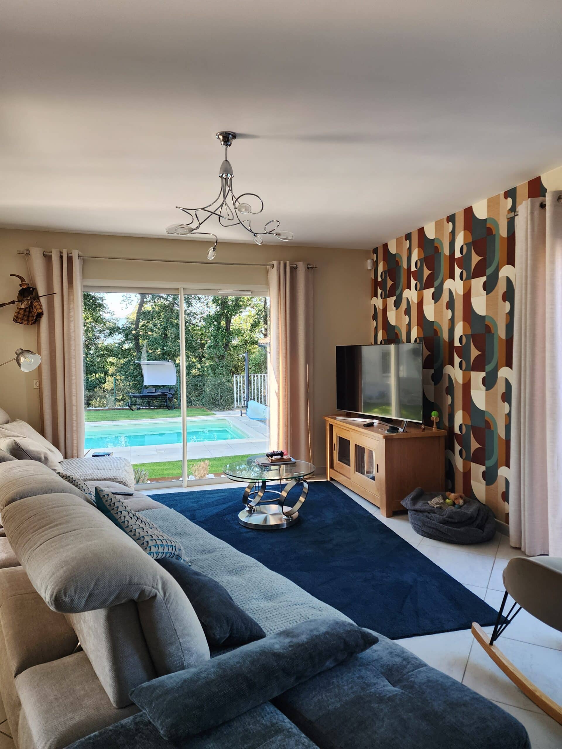 Salon moderne avec canapé gris, mur géométrique et baie vitrée ouvrant sur une piscine ensoleillée.
