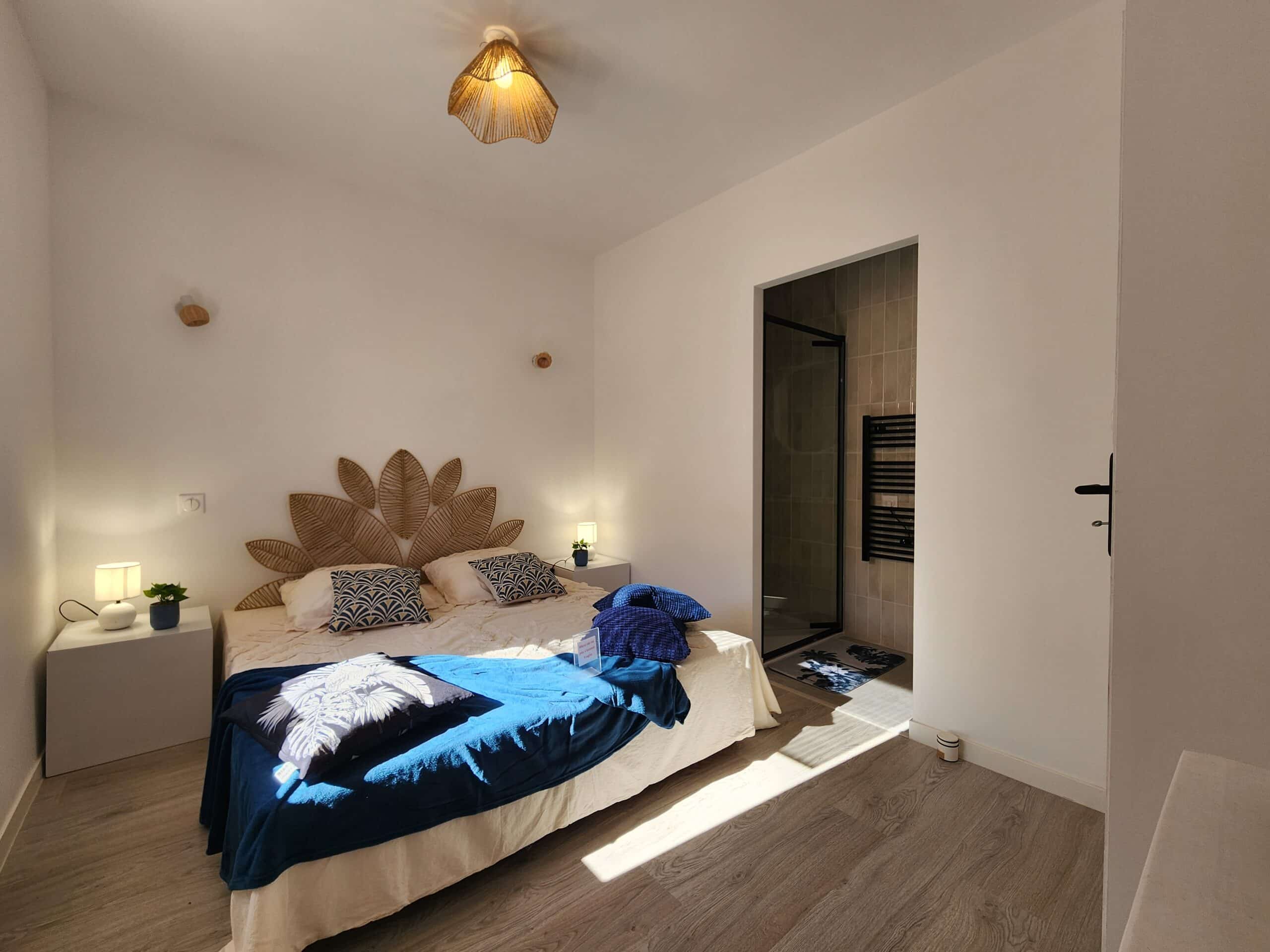 Chambre d'hôte lumineuse : grand lit, tête de lit tressée en feuille. Couverture bleue (avec l'étiquette "Fragile"). Douche moderne attenante.