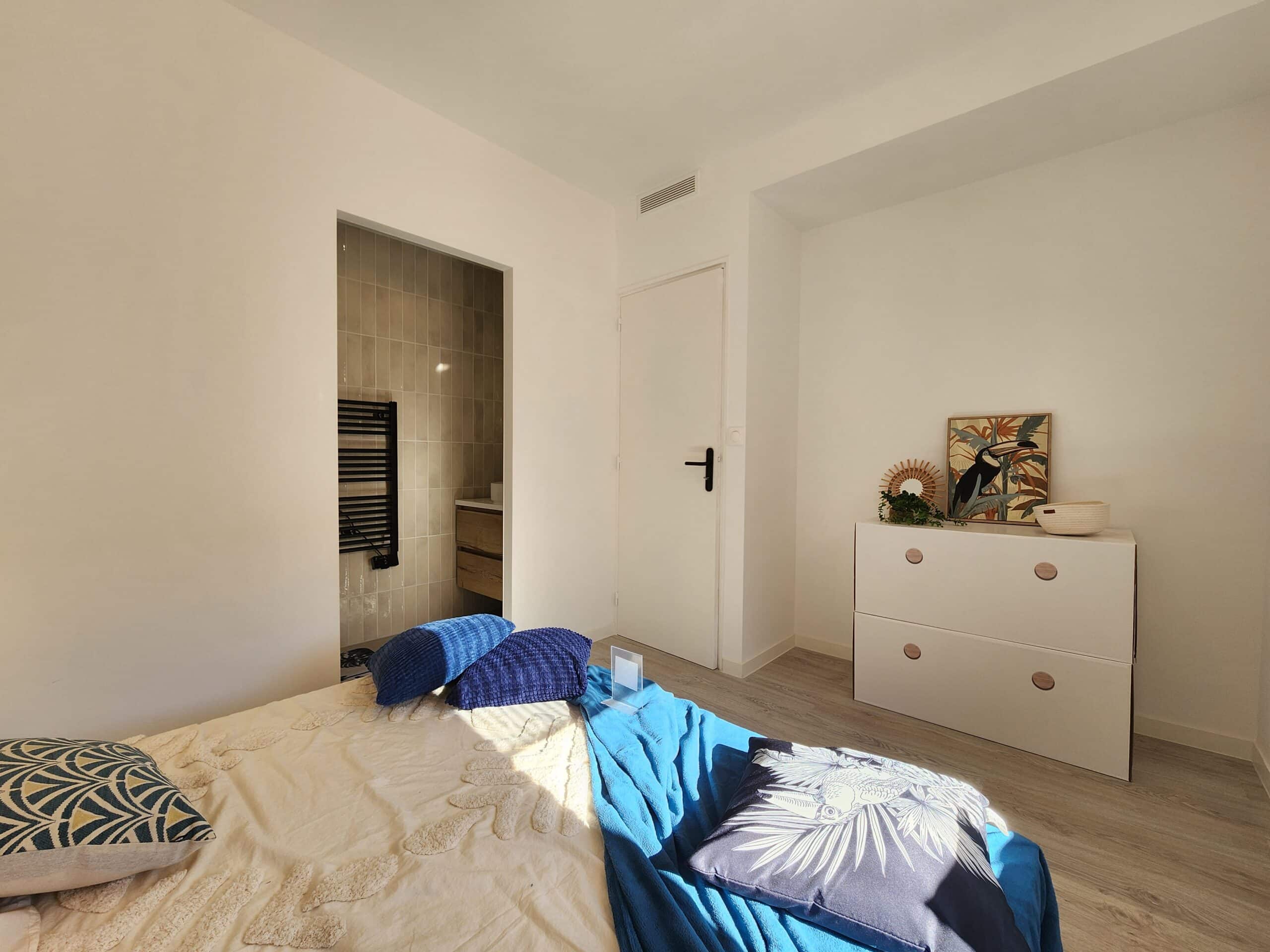 Chambre moderne lumineuse. Lit, jeté bleu et coussins. Commode blanche avec tableau Toucan tropical. Entrée salle de bain carrelée.