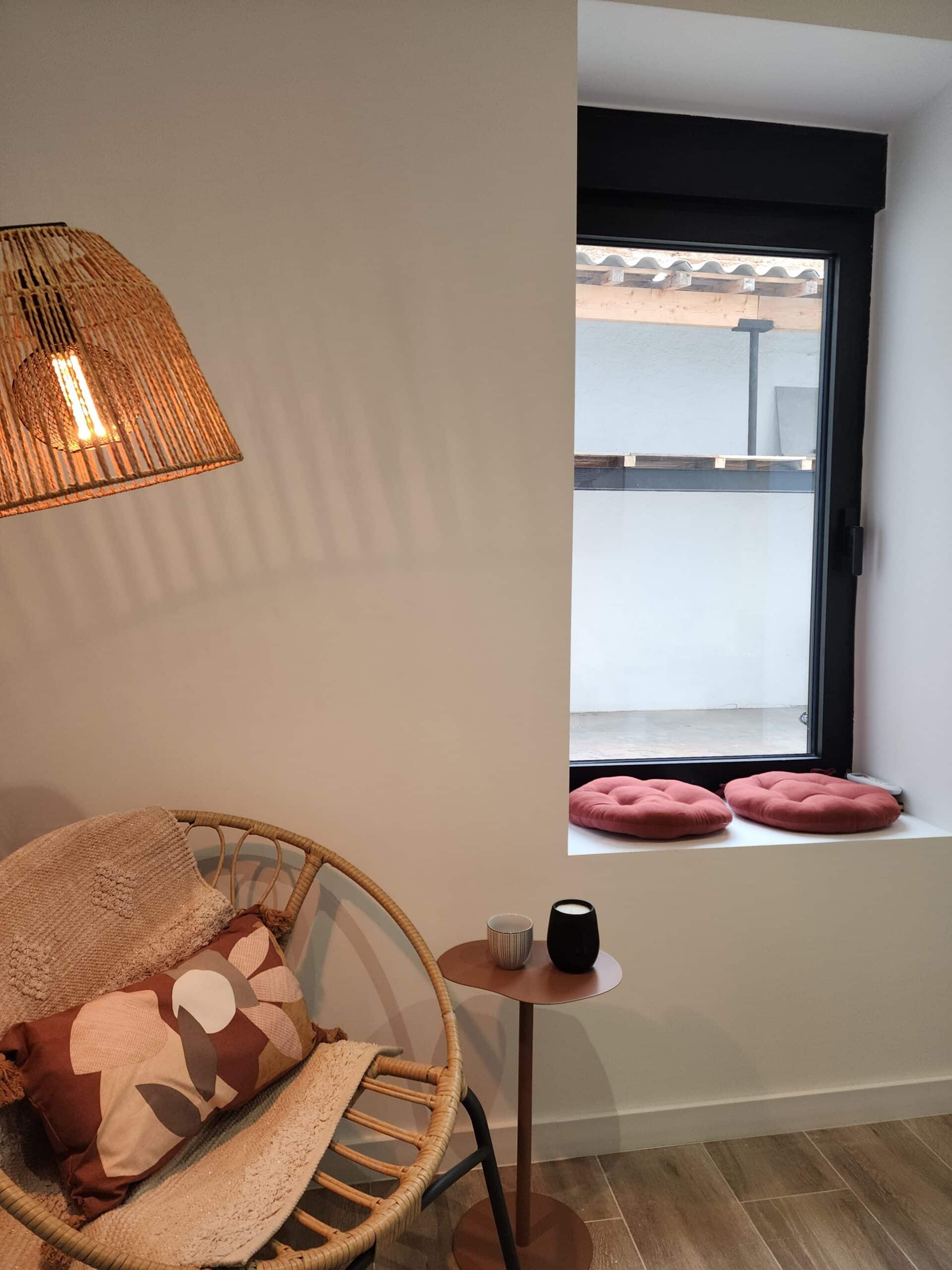 Fauteuil en rotin et couverture douillette dans un coin lecture. Lampe en jute, table d'appoint, et coussins ronds terracotta près de la fenêtre.