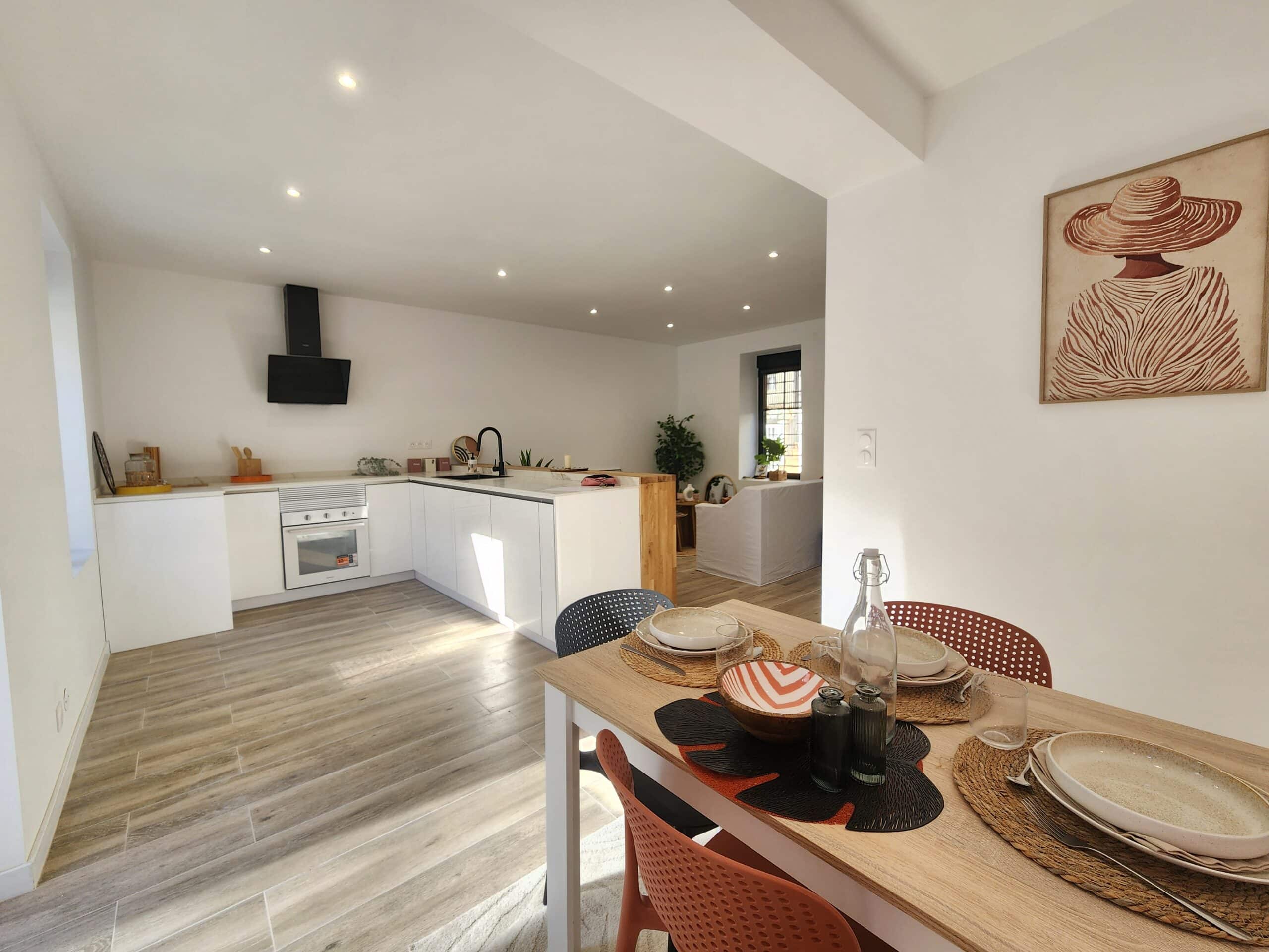 Salle à manger moderne avec art mural face à une cuisine ouverte blanche, îlot central et robinet noir. Carrelage effet bois.