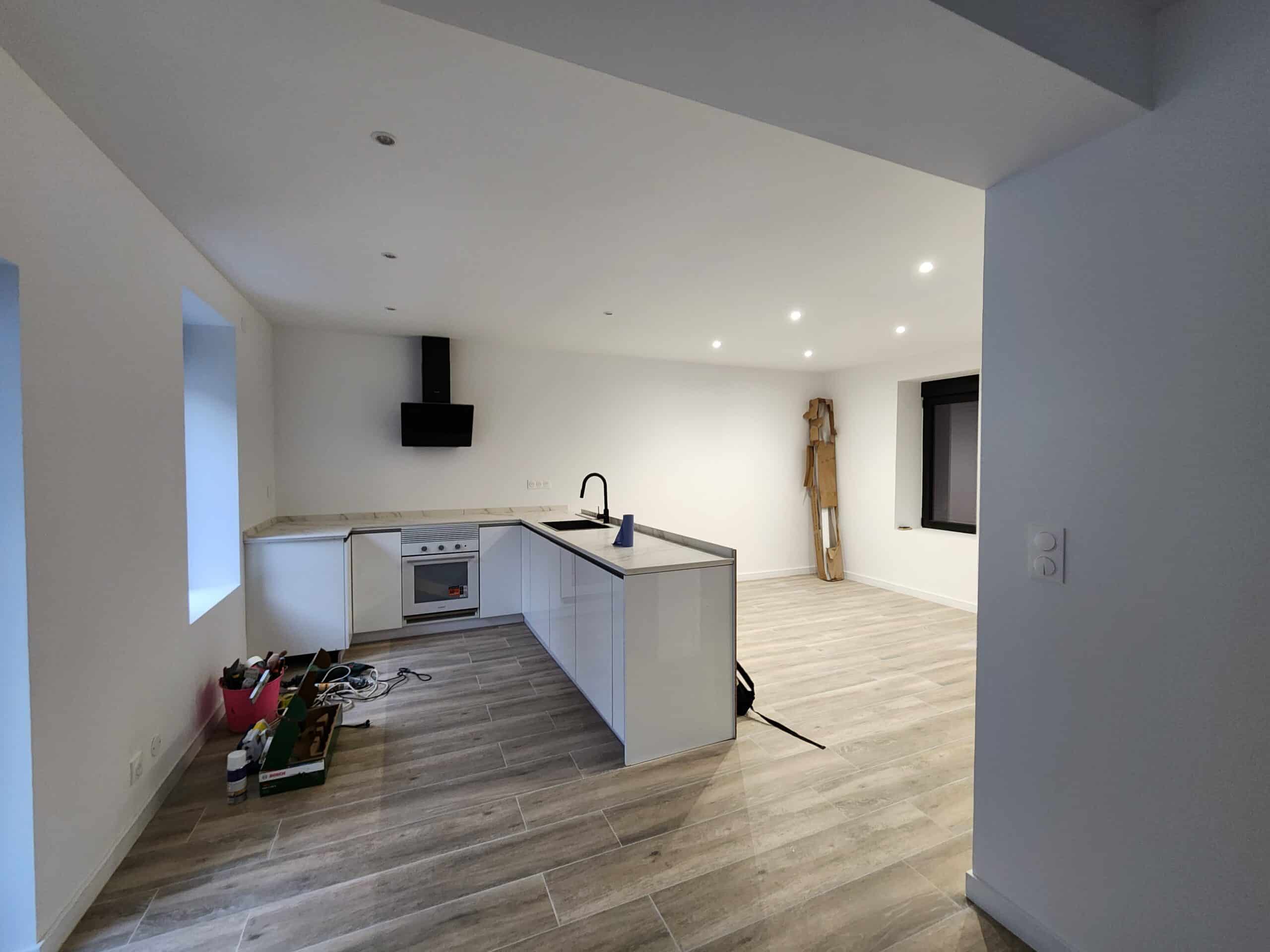 Cuisine ouverte moderne en L avec armoires blanches laquées, hotte noire et comptoir marbré, sur sol carrelé aspect bois. Pièce en fin de rénovation.