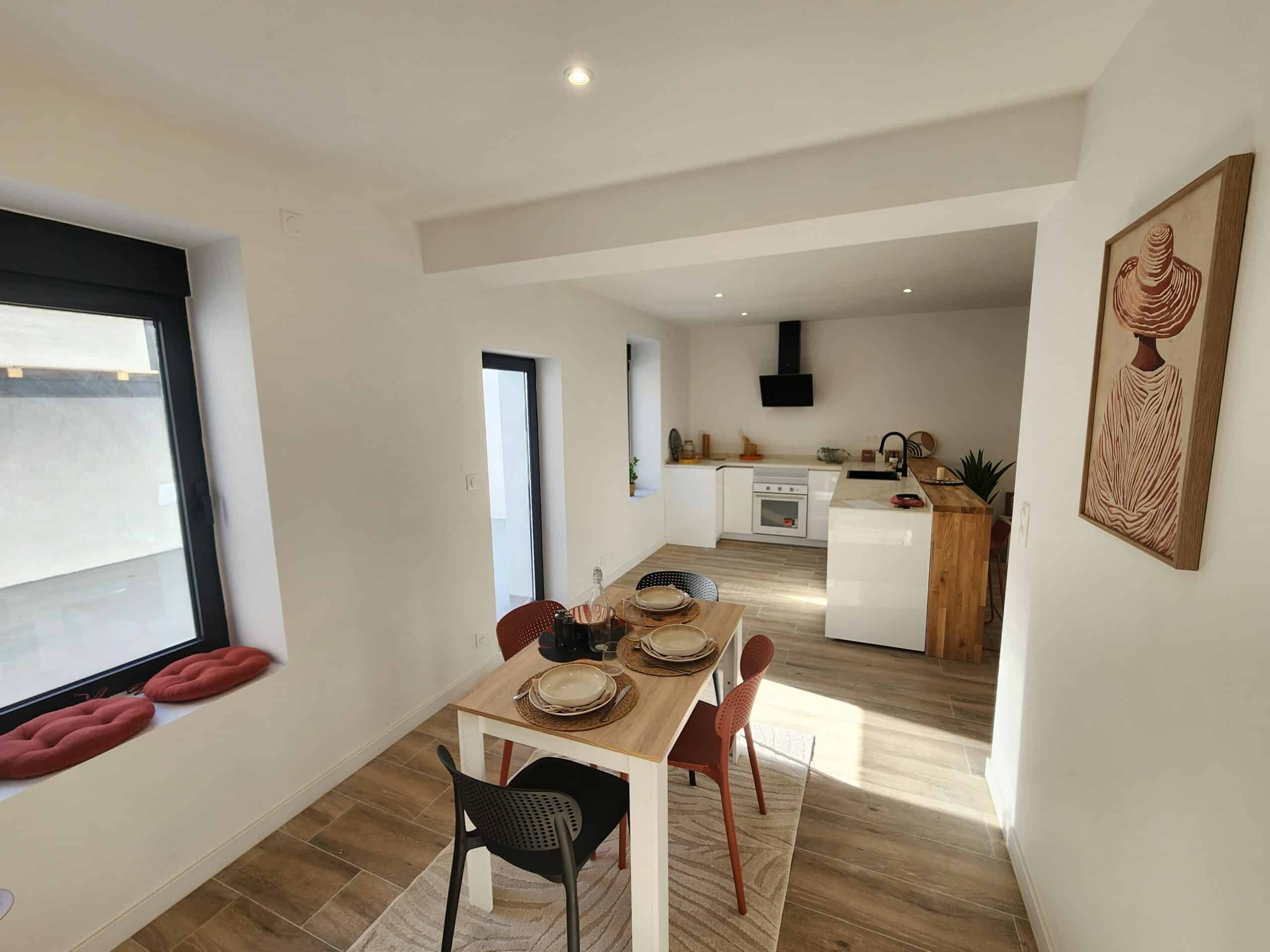 Cuisine ouverte lumineuse et salle à manger minimaliste. Mobilier blanc laqué, table en bois clair, carrelage imitation bois, et art mural.