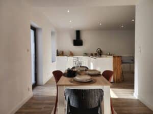 Salle à manger moderne et cuisine ouverte blanche. Table dressée, chaises design noires et rouges. Îlot central. Spots LED. Pots "Pâtes, Riz, Farine."