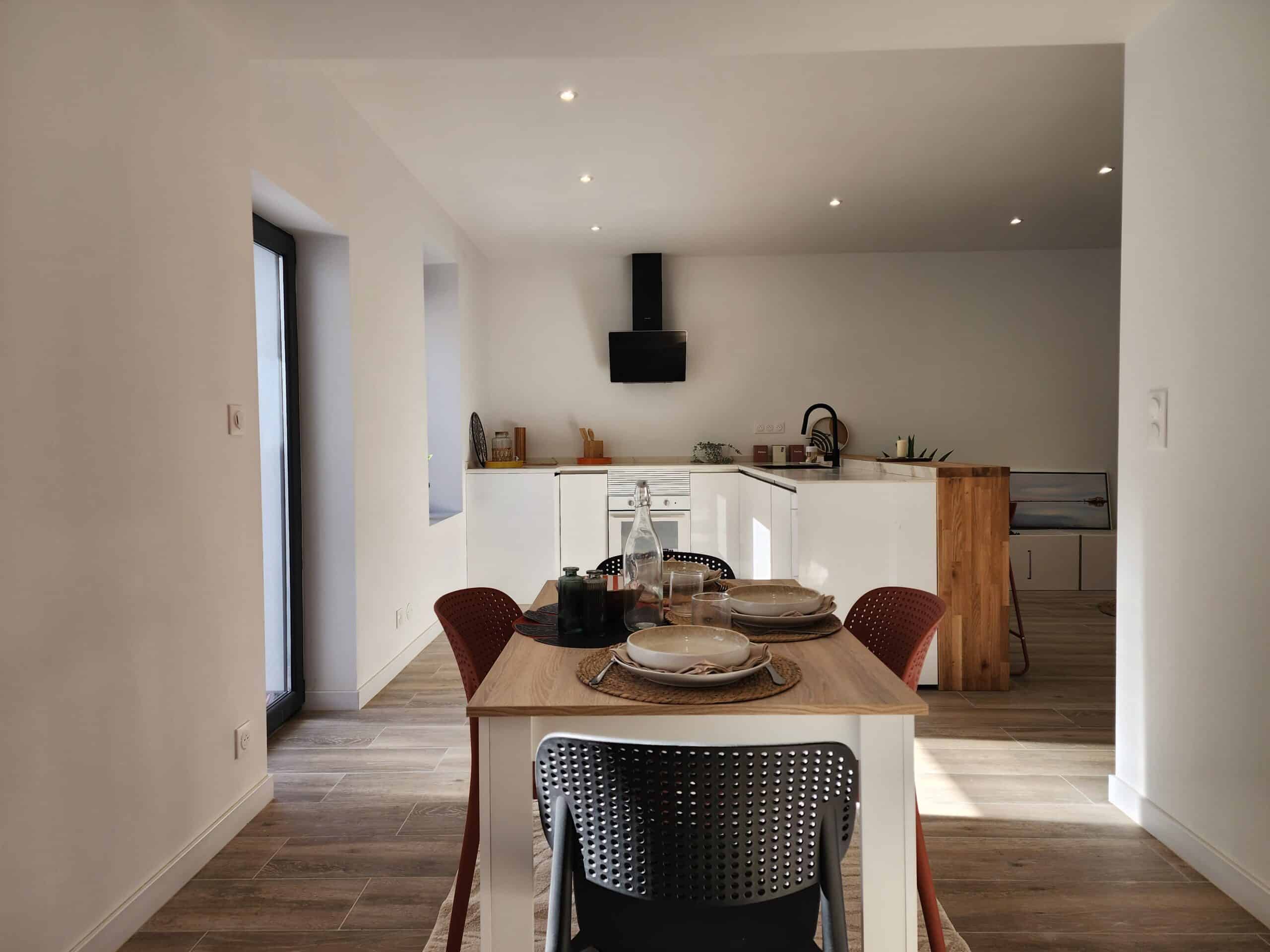 Salle à manger moderne et cuisine ouverte blanche. Table dressée, chaises design noires et rouges. Îlot central. Spots LED. Pots "Pâtes, Riz, Farine."