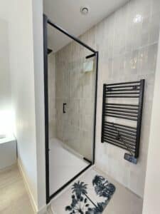 Douche italienne moderne à cadre noir et receveur blanc. Carrelage beige vertical brillant, sèche-serviettes noir Equation fixé au mur.