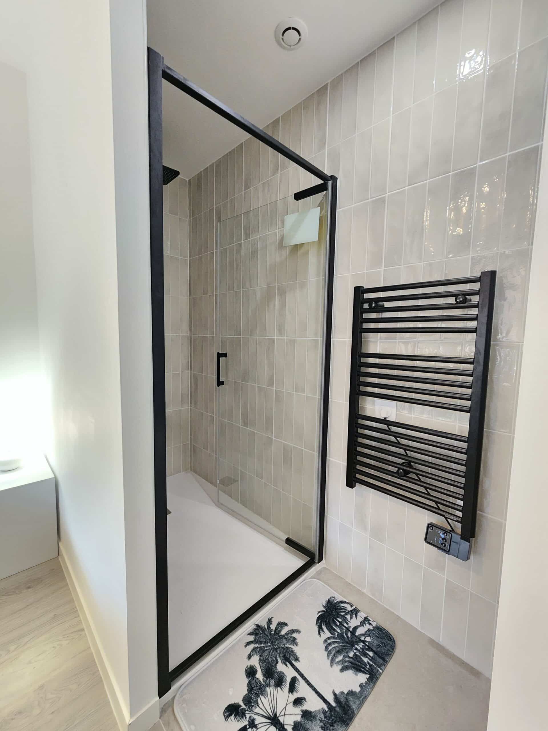 Douche italienne moderne à cadre noir et receveur blanc. Carrelage beige vertical brillant, sèche-serviettes noir Equation fixé au mur.