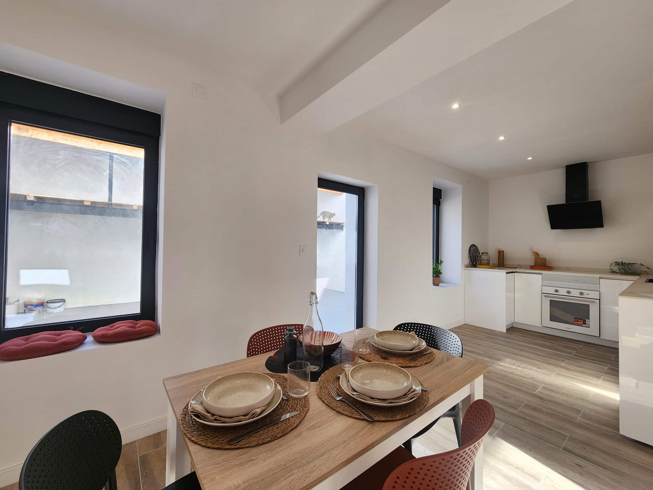 Intérieur moderne et lumineux. Cuisine ouverte blanche, hotte noire, et salle à manger avec table en bois dressée pour quatre personnes.