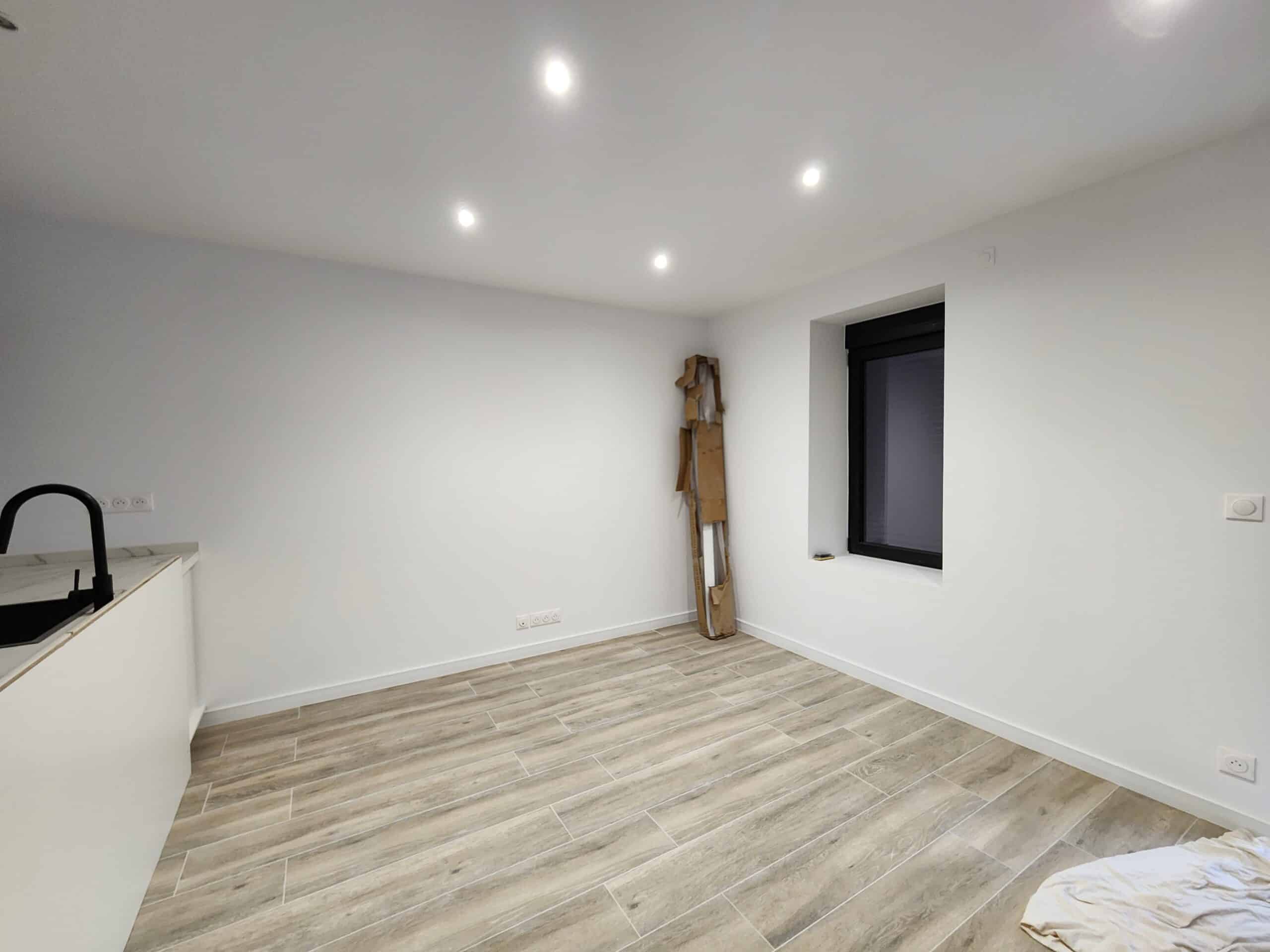 Pièce rénovée style moderne. Murs blancs, spots, et carrelage au sol aspect bois. Cuisine minimaliste et fenêtre encadrée de noir.