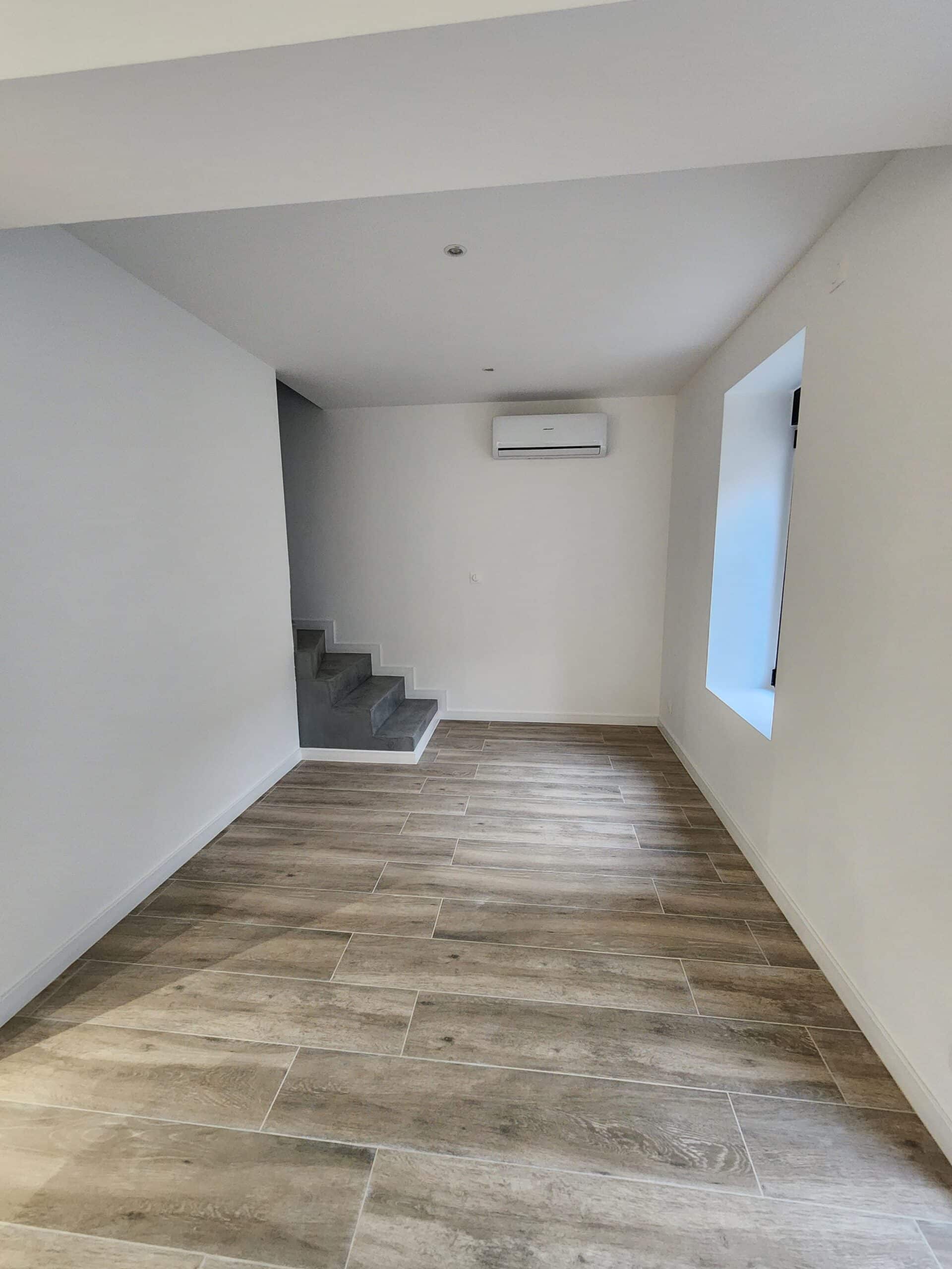 Intérieur moderne, murs blancs, escalier en béton gris, carrelage imitation bois clair. Climatiseur mural et petite fenêtre.