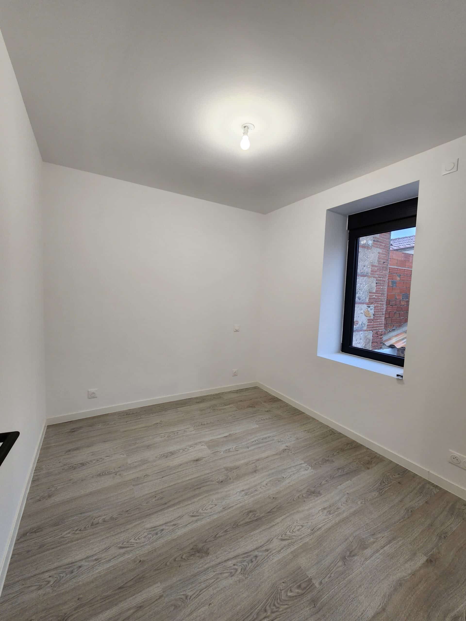 Pièce vide rénovée avec murs blancs et sol stratifié gris clair. Fenêtre noire encastrée et éclairage minimaliste au plafond.