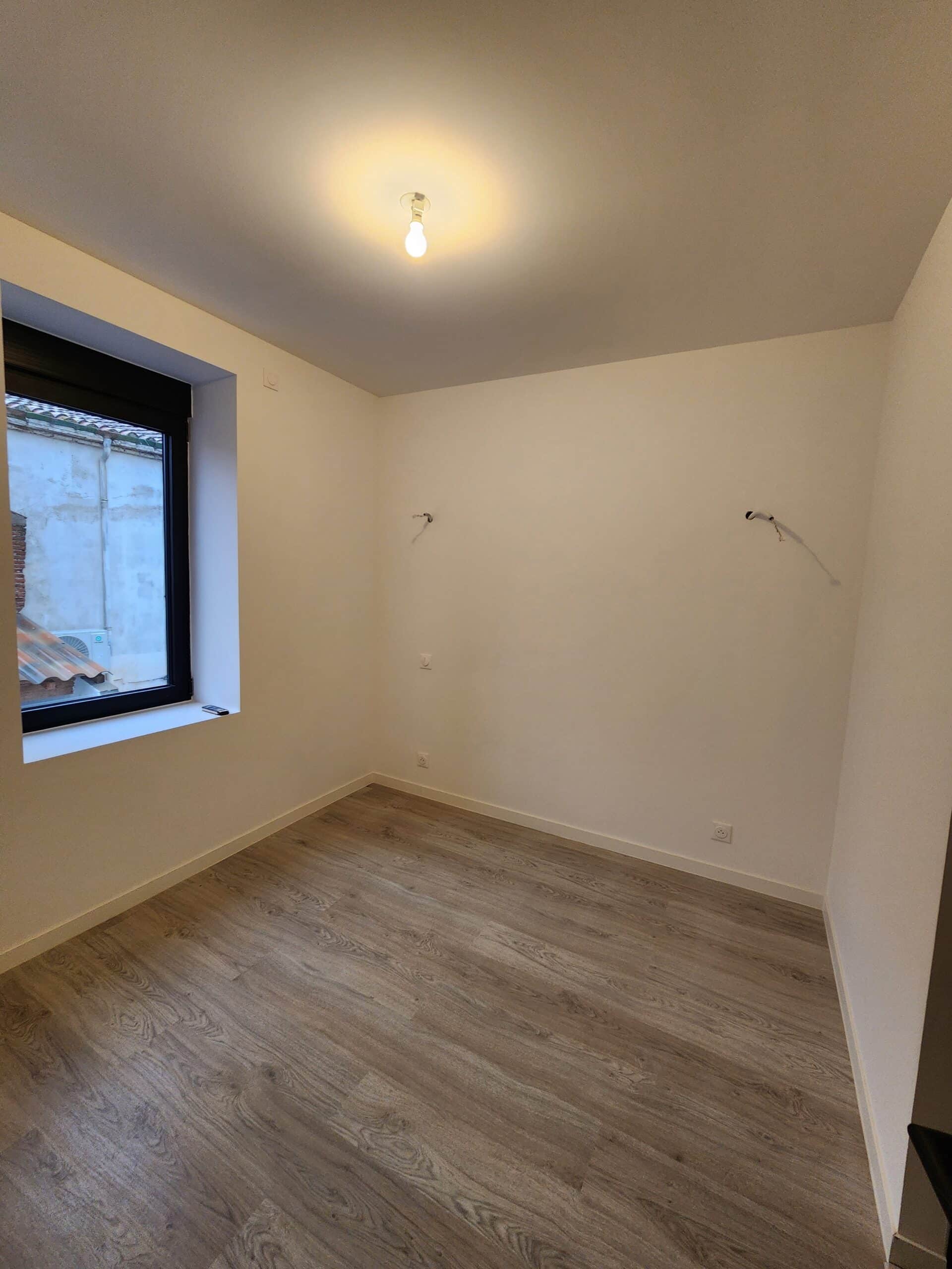 Chambre vide et lumineuse avec murs blancs, sol stratifié bois, et câbles en attente. Une fenêtre noire donne sur une cour intérieure.