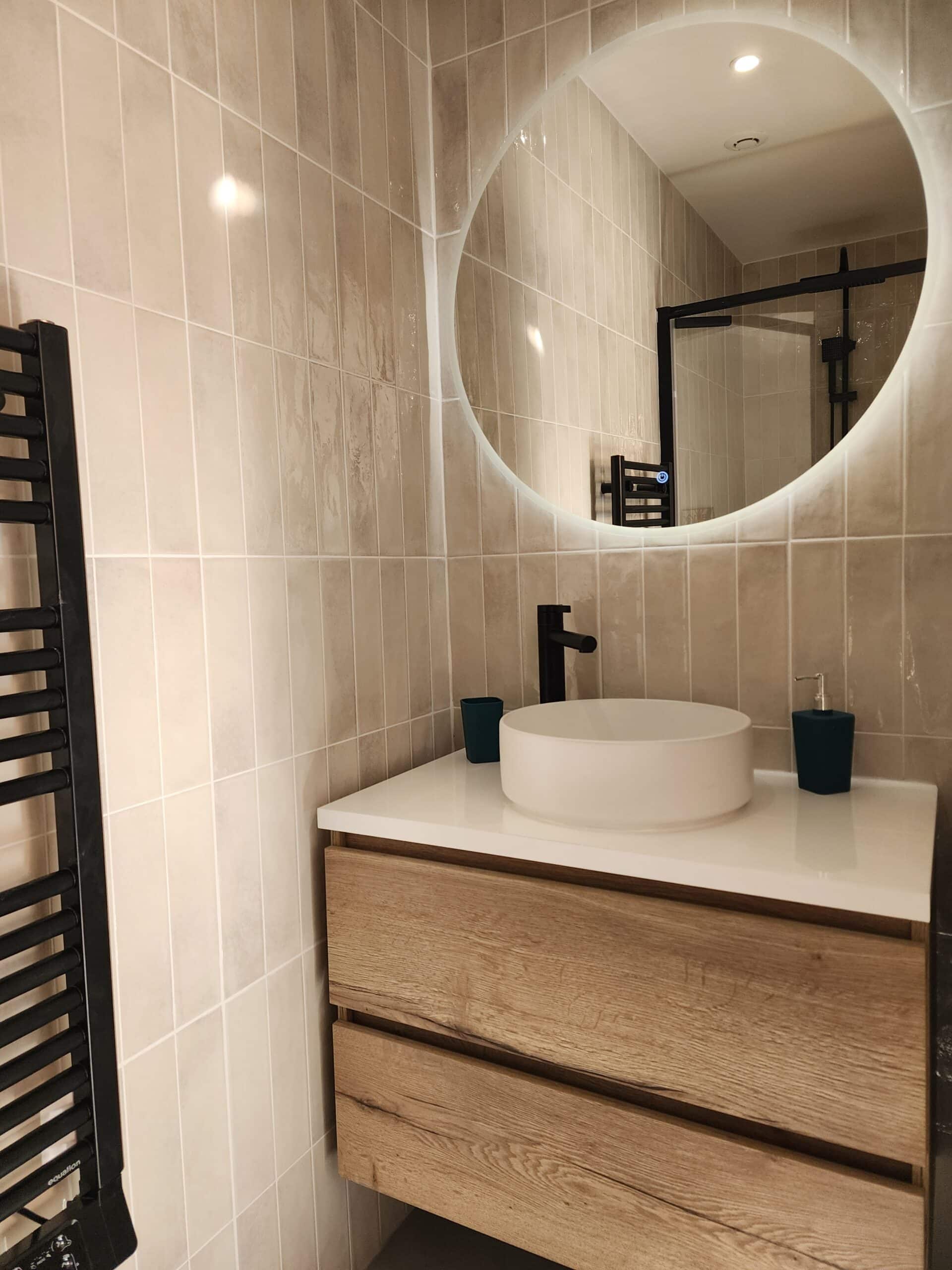 Meuble vasque bois, vasque blanche, robinetterie et sèche-serviettes noirs. Grand miroir rond LED sur mur de carrelage beige vertical.