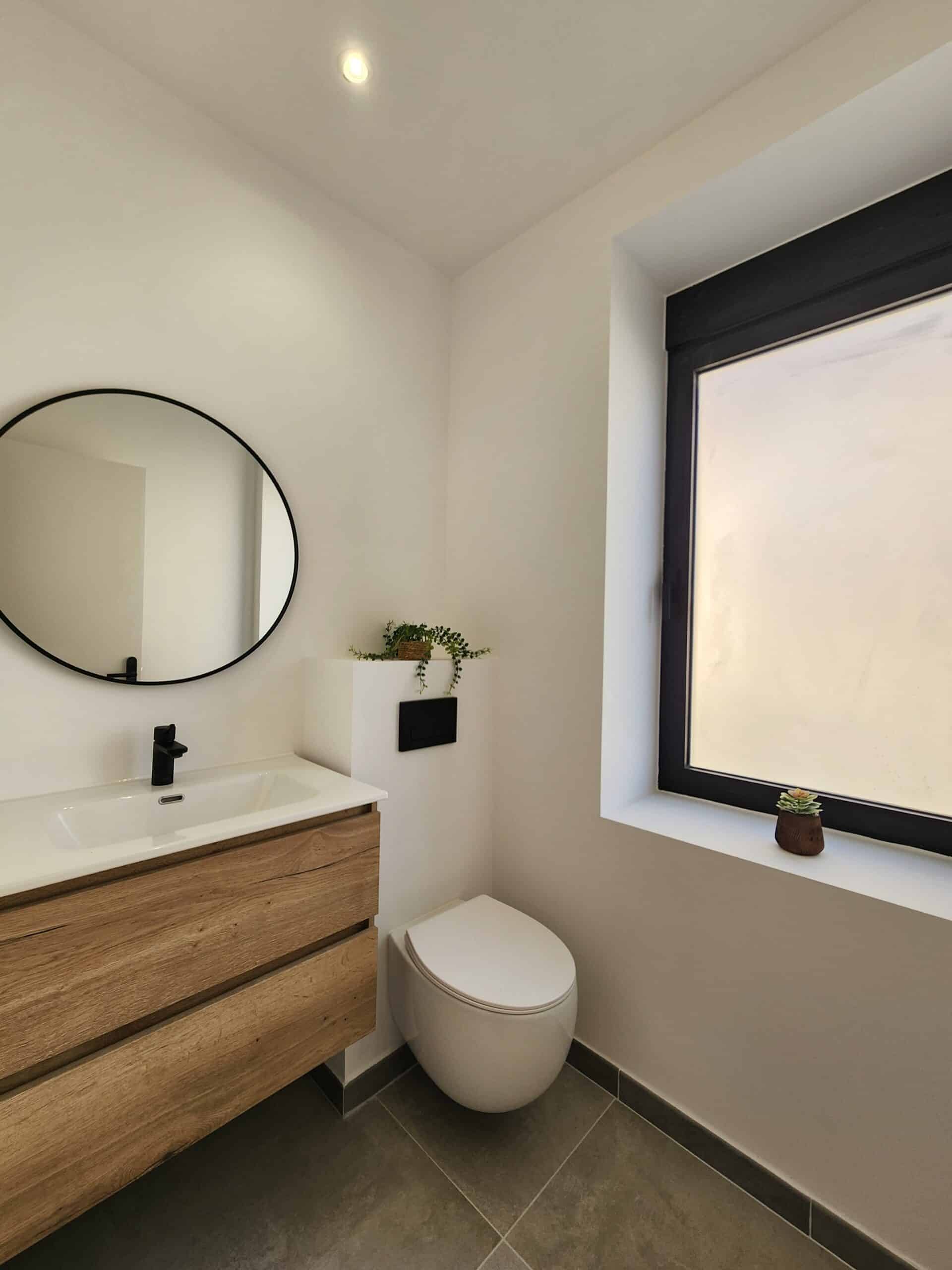 Salle d'eau moderne : meuble bois flottant, vasque et robinet noirs. Miroir rond, WC suspendu, murs blancs et carrelage gris.