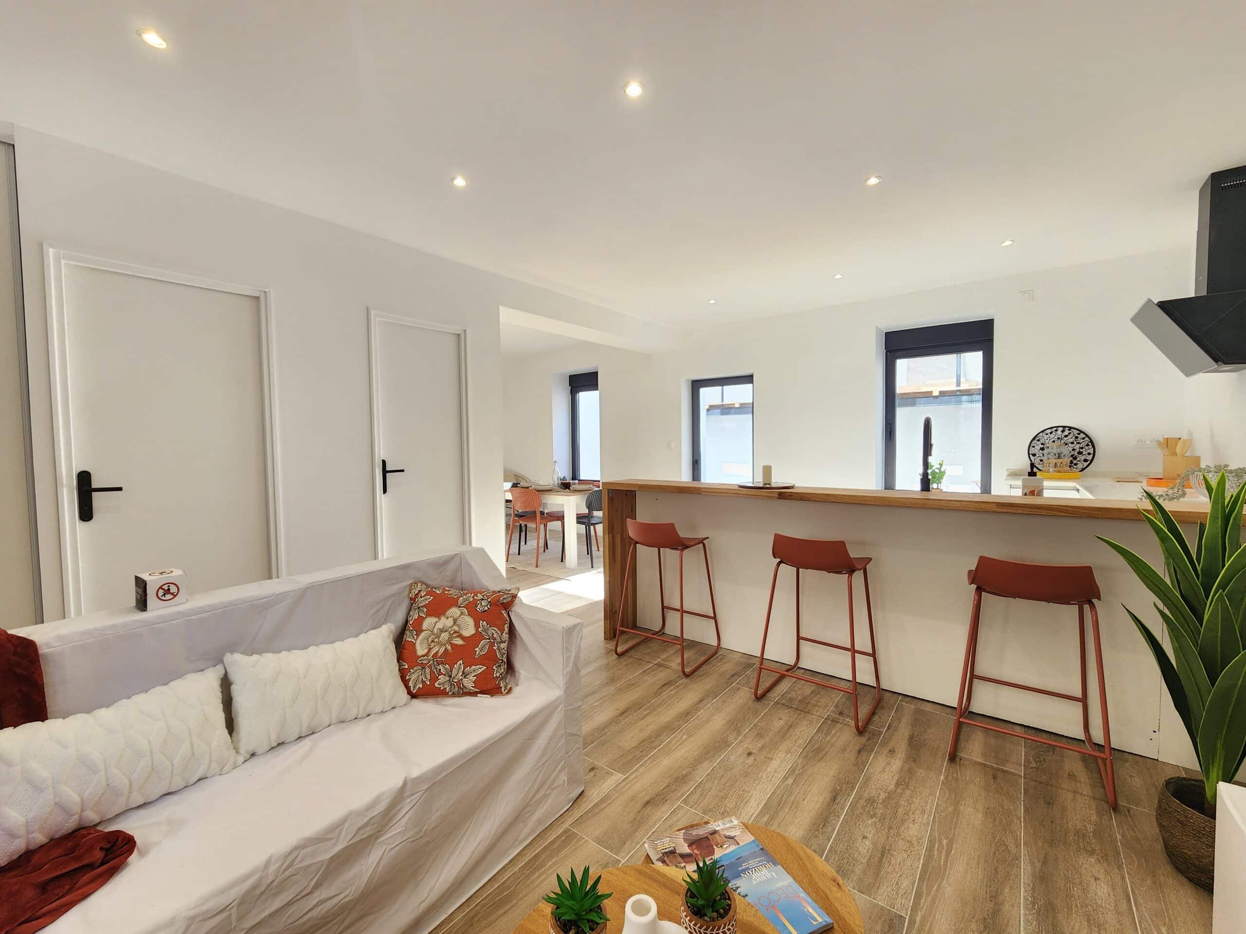 Intérieur moderne. Salon ouvert sur cuisine avec comptoir bar en bois et trois tabourets rouges. Canapé blanc, portes intérieures et salle à manger en arrière-plan.