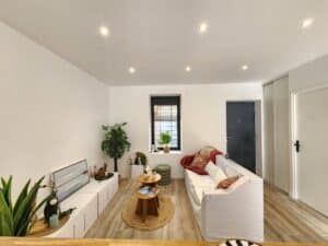 Salon moderne lumineux, murs blancs. Canapé blanc, tables basses en bois sur tapis en jute. Décoration naturelle et plantes. Sol en carrelage imitation bois.