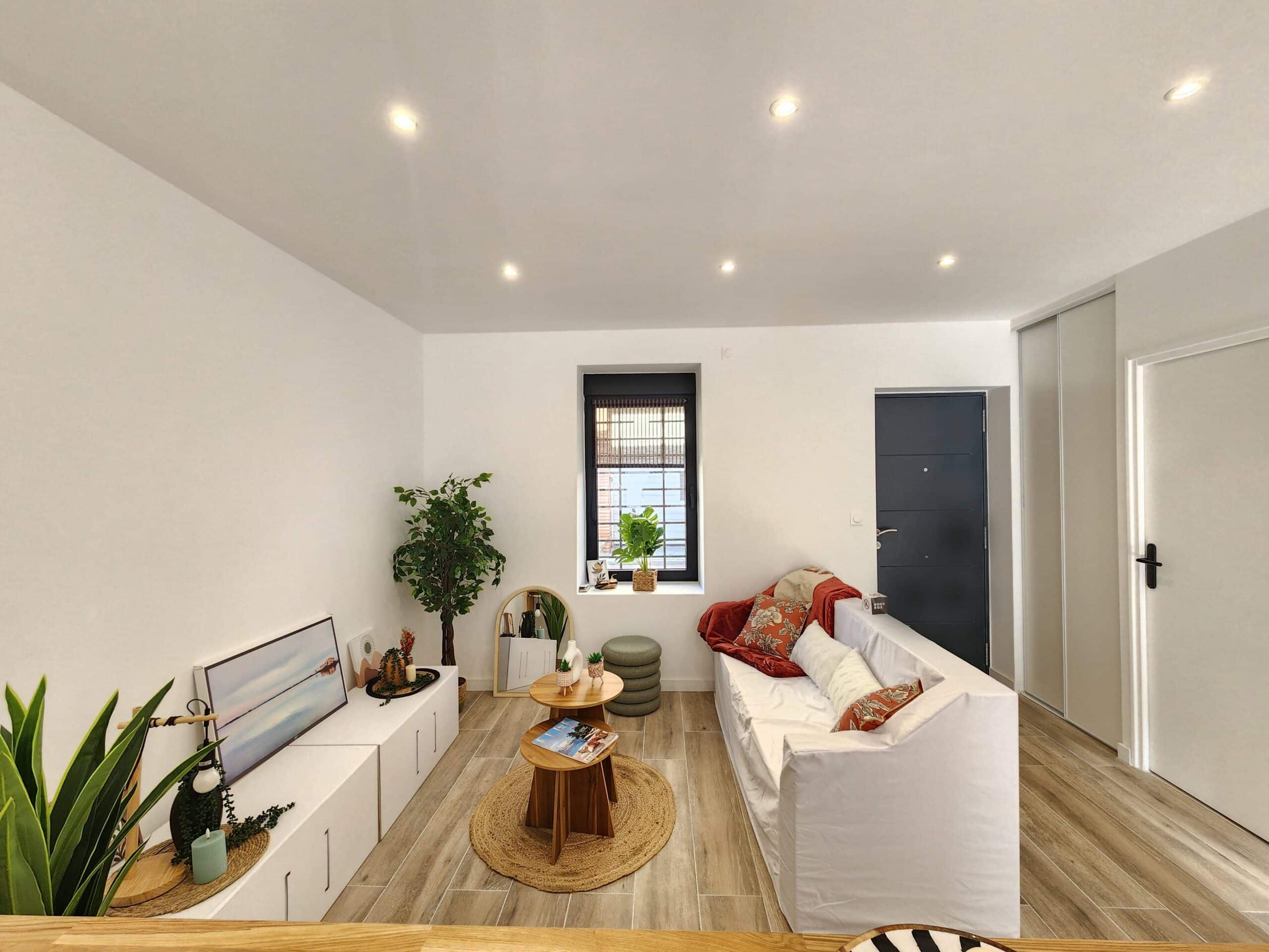 Salon moderne lumineux, murs blancs. Canapé blanc, tables basses en bois sur tapis en jute. Décoration naturelle et plantes. Sol en carrelage imitation bois.