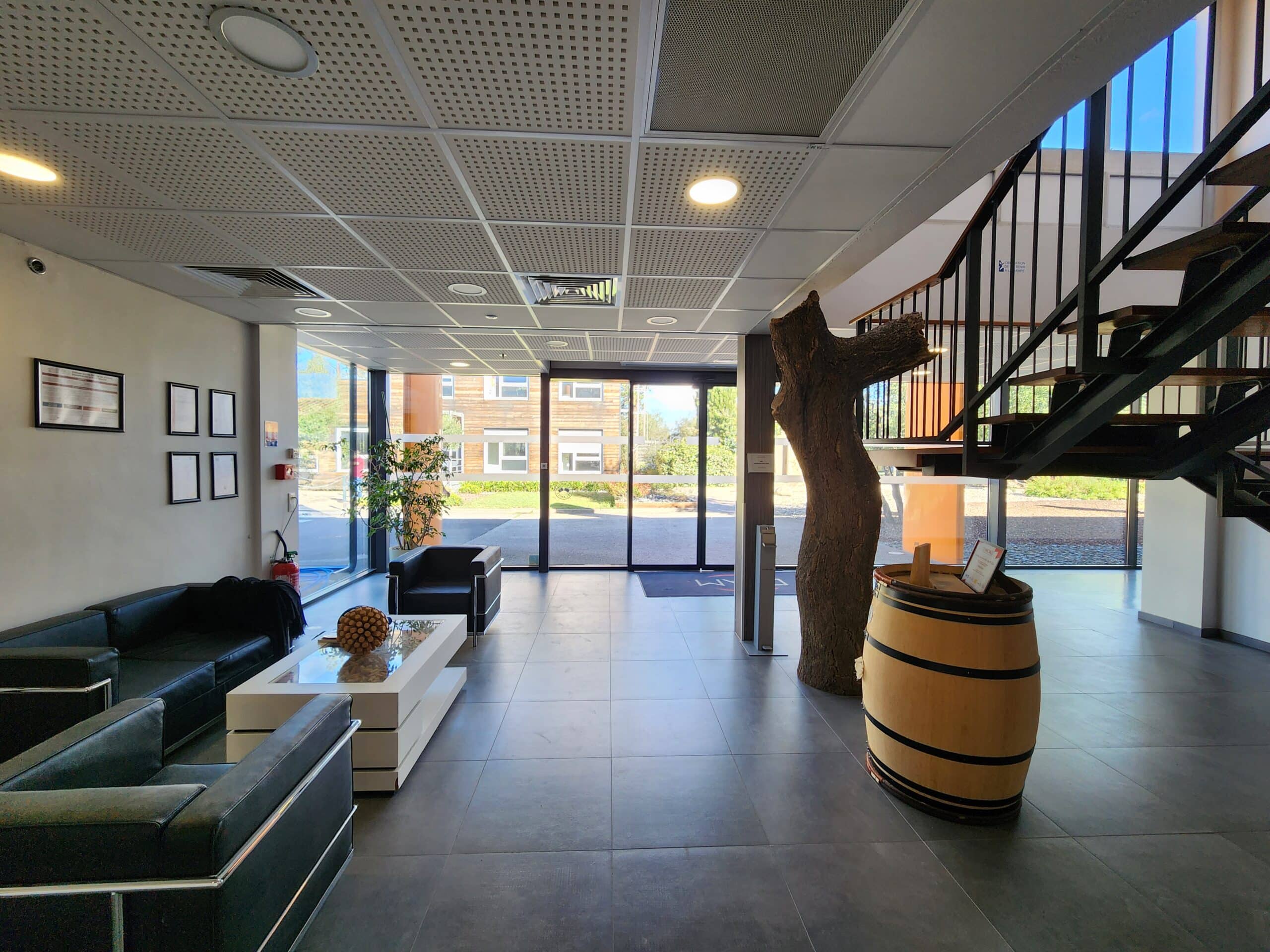 Lobby moderne avec canapés noirs, table blanche, baril de vin et sculpture d'arbre près d'une baie vitrée.