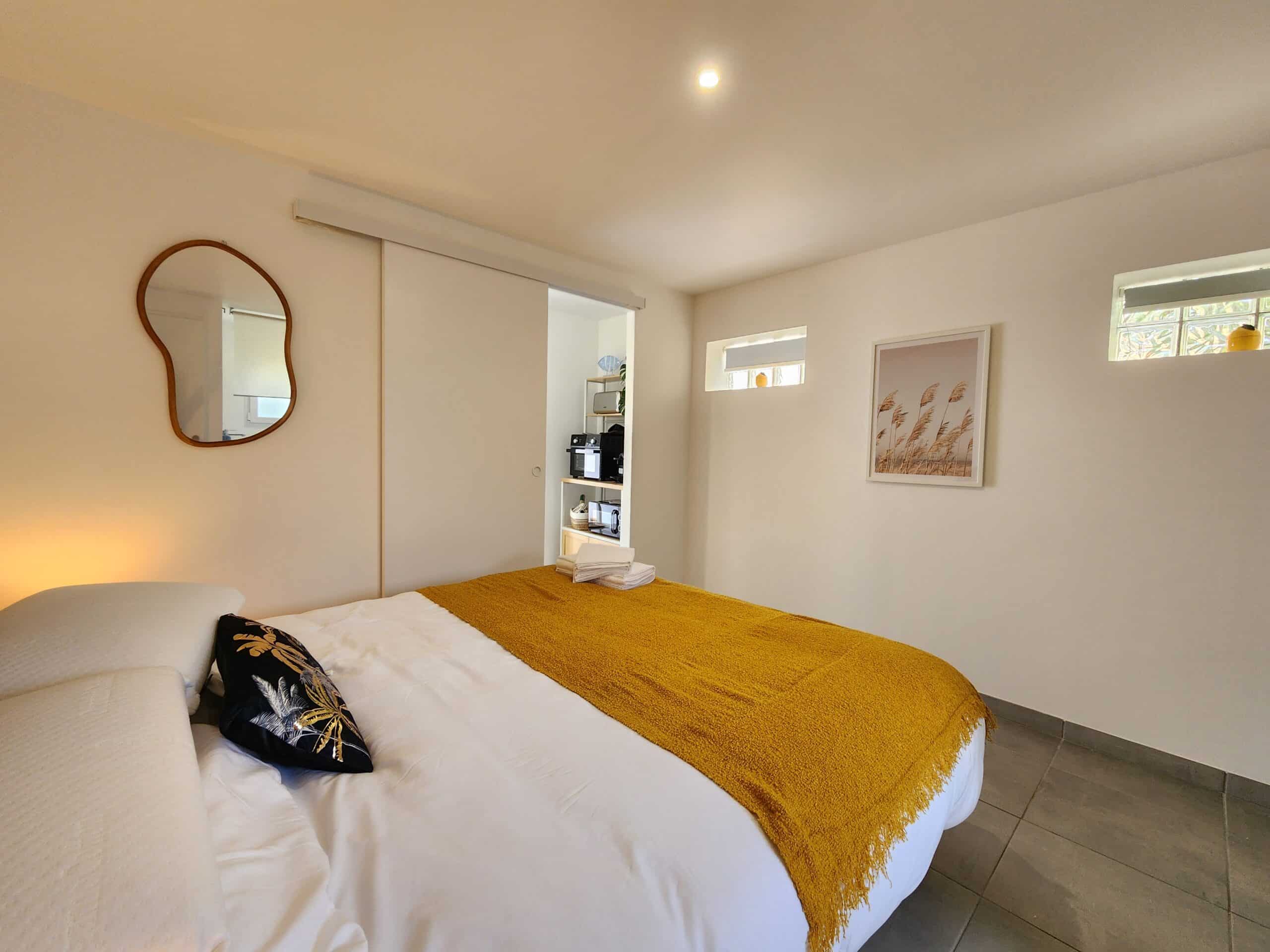 Chambre moderne, lit avec jeté moutarde, miroir design et porte coulissante ouvrant sur une kitchenette.