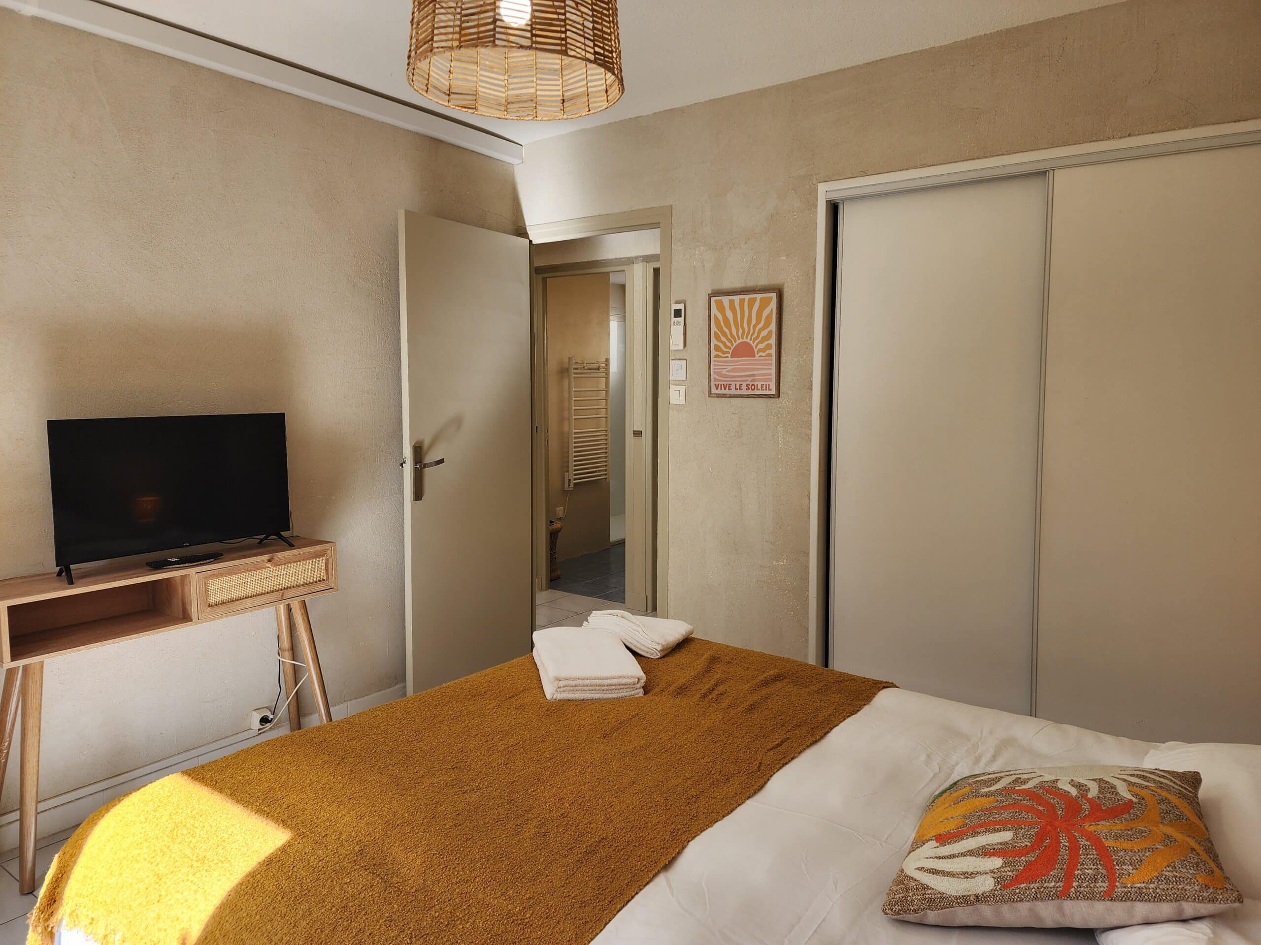 Chambre avec lit jeté moutarde, meuble TV bois et affiche 'VIVE LE SOLEIL'.