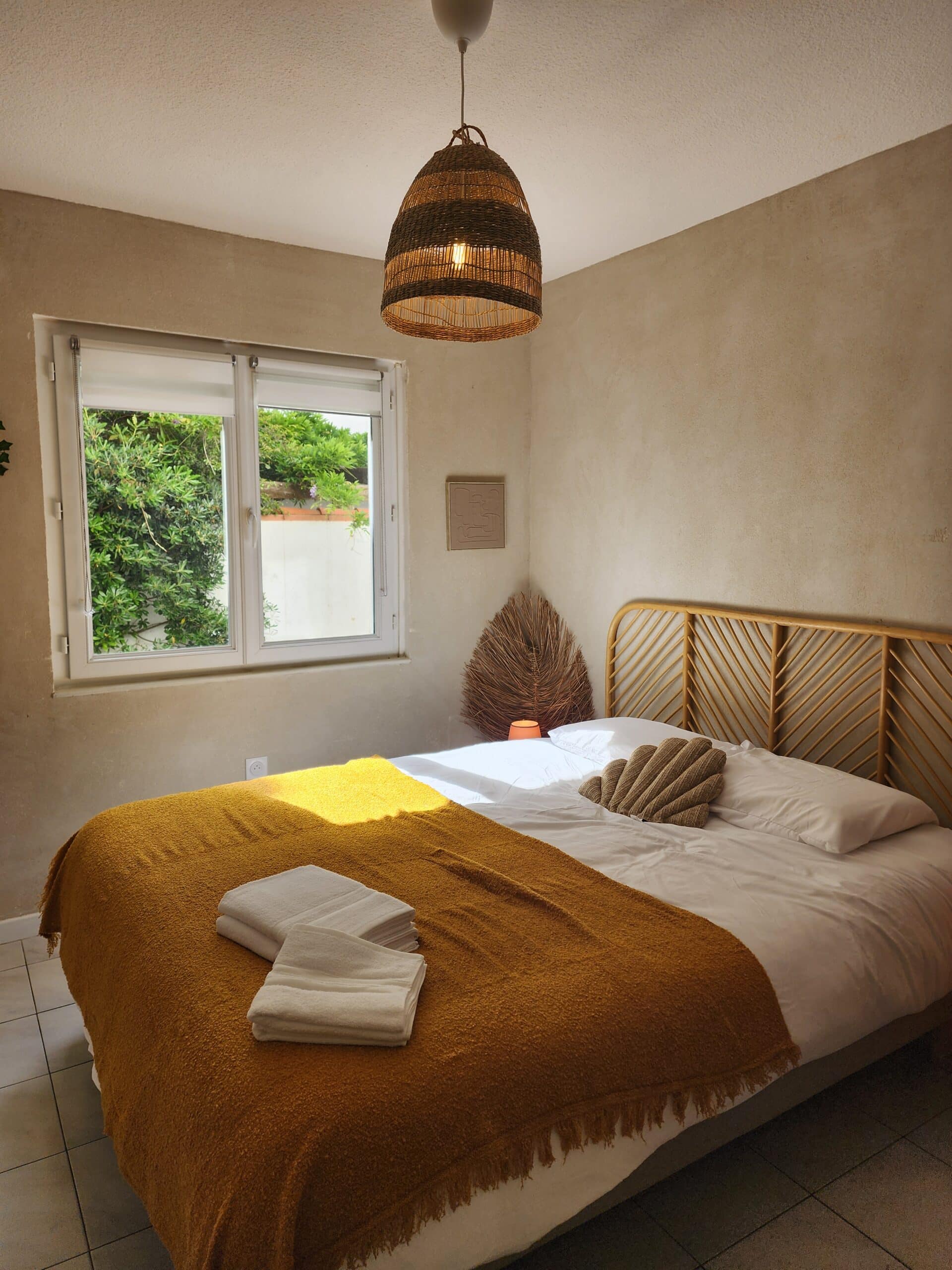 Chambre bohème avec lit en rotin, jeté de lit moutarde et luminaire tressé devant une fenêtre ensoleillée.