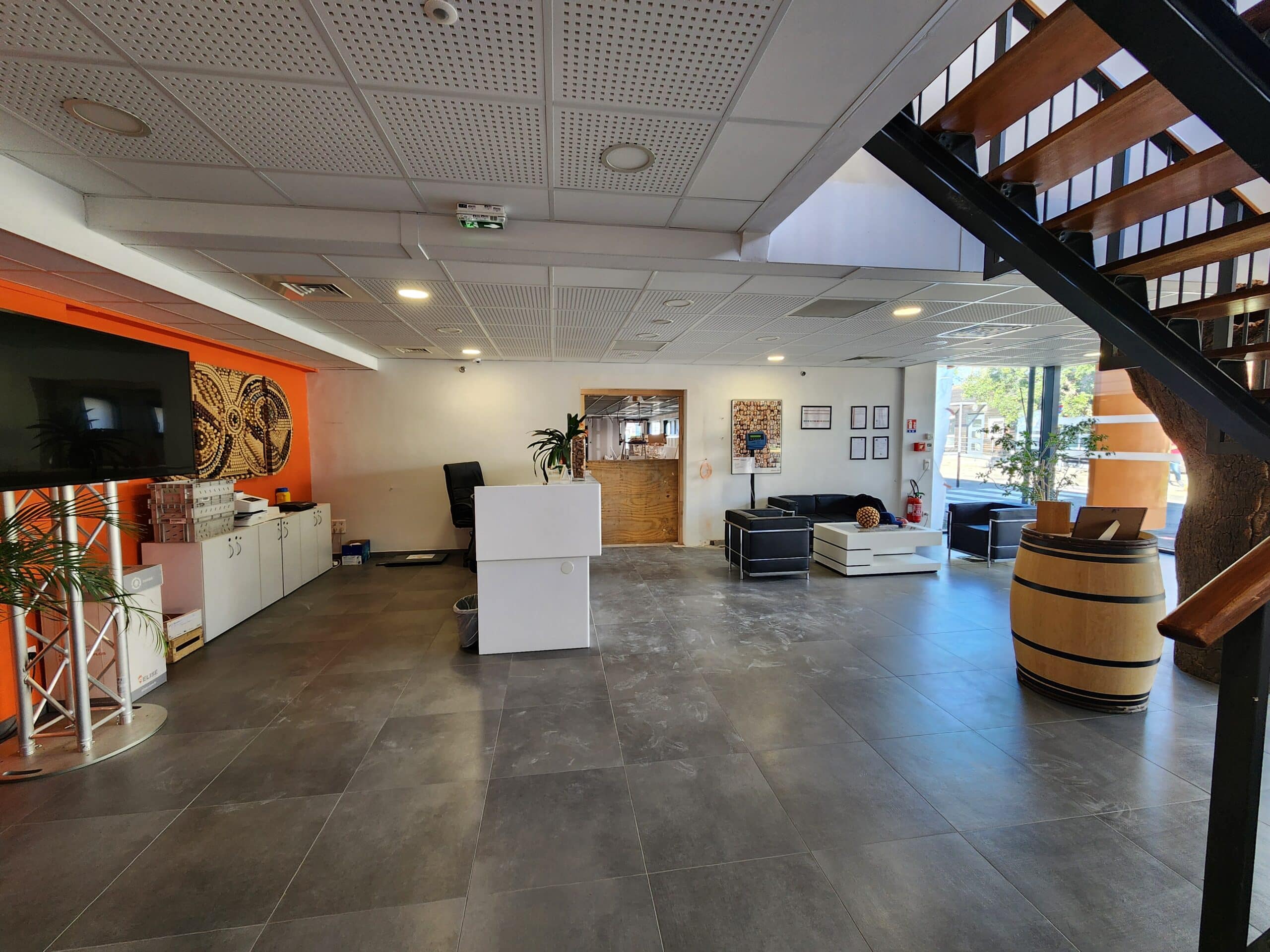 Hall d'accueil moderne : bureau blanc, salon noir, barrique décorative et escalier industriel sur sol gris.