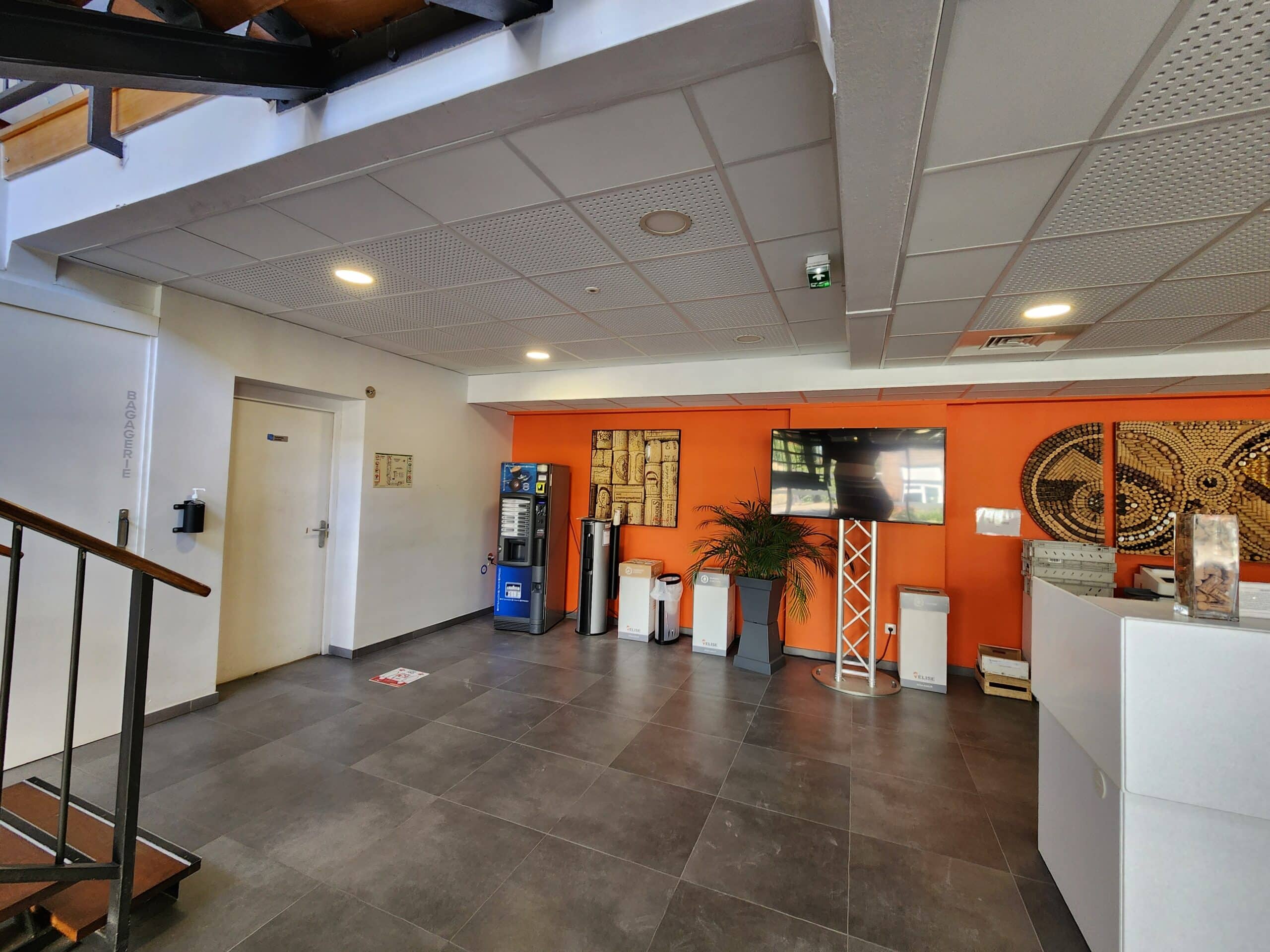 Espace d'accueil avec mur orange, machine à café Lavazza, écran TV et accès bagagerie.