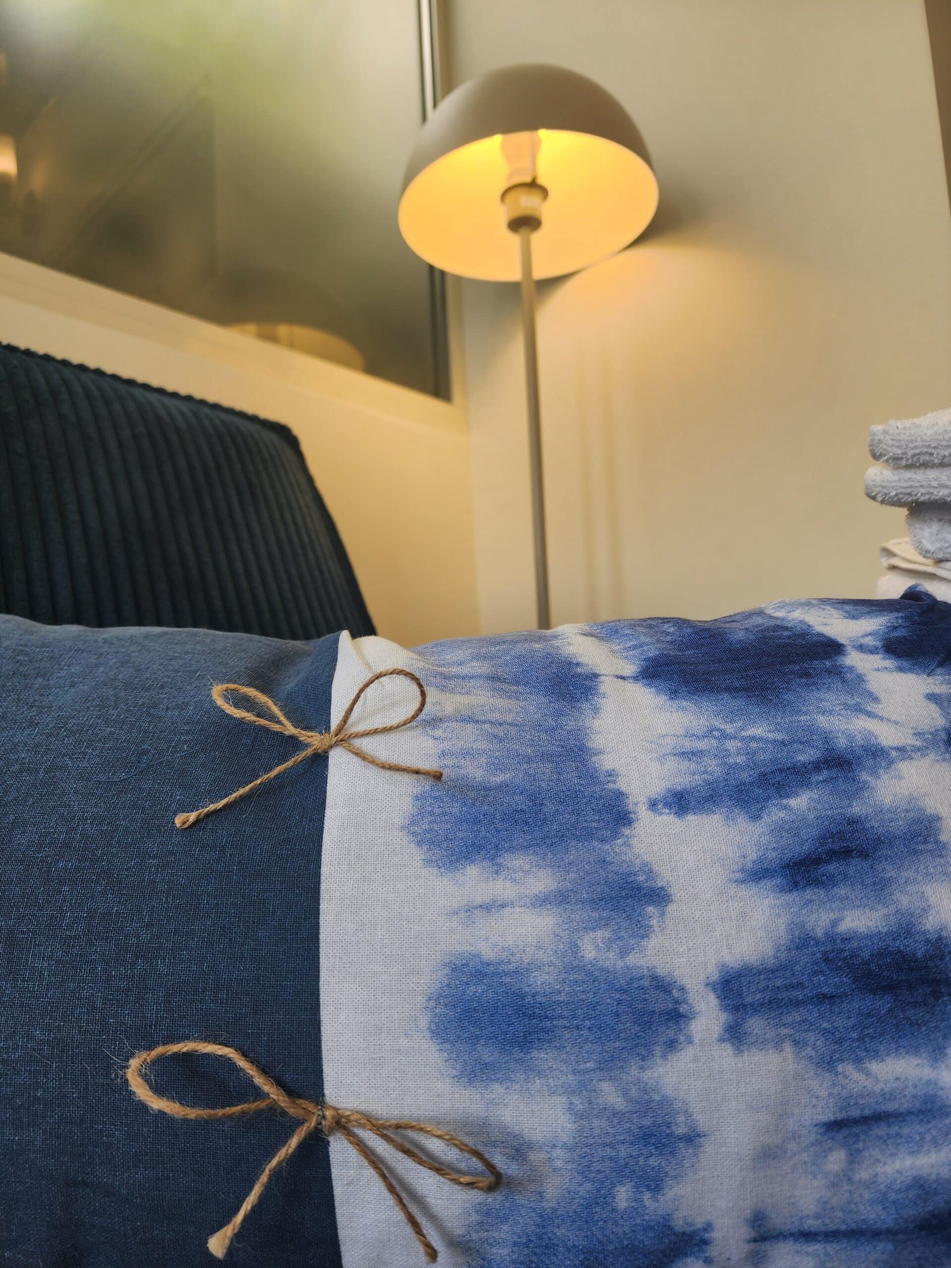 Oreiller bleu tie-dye avec nœuds en ficelle devant une lampe design allumée dans une chambre chaleureuse.