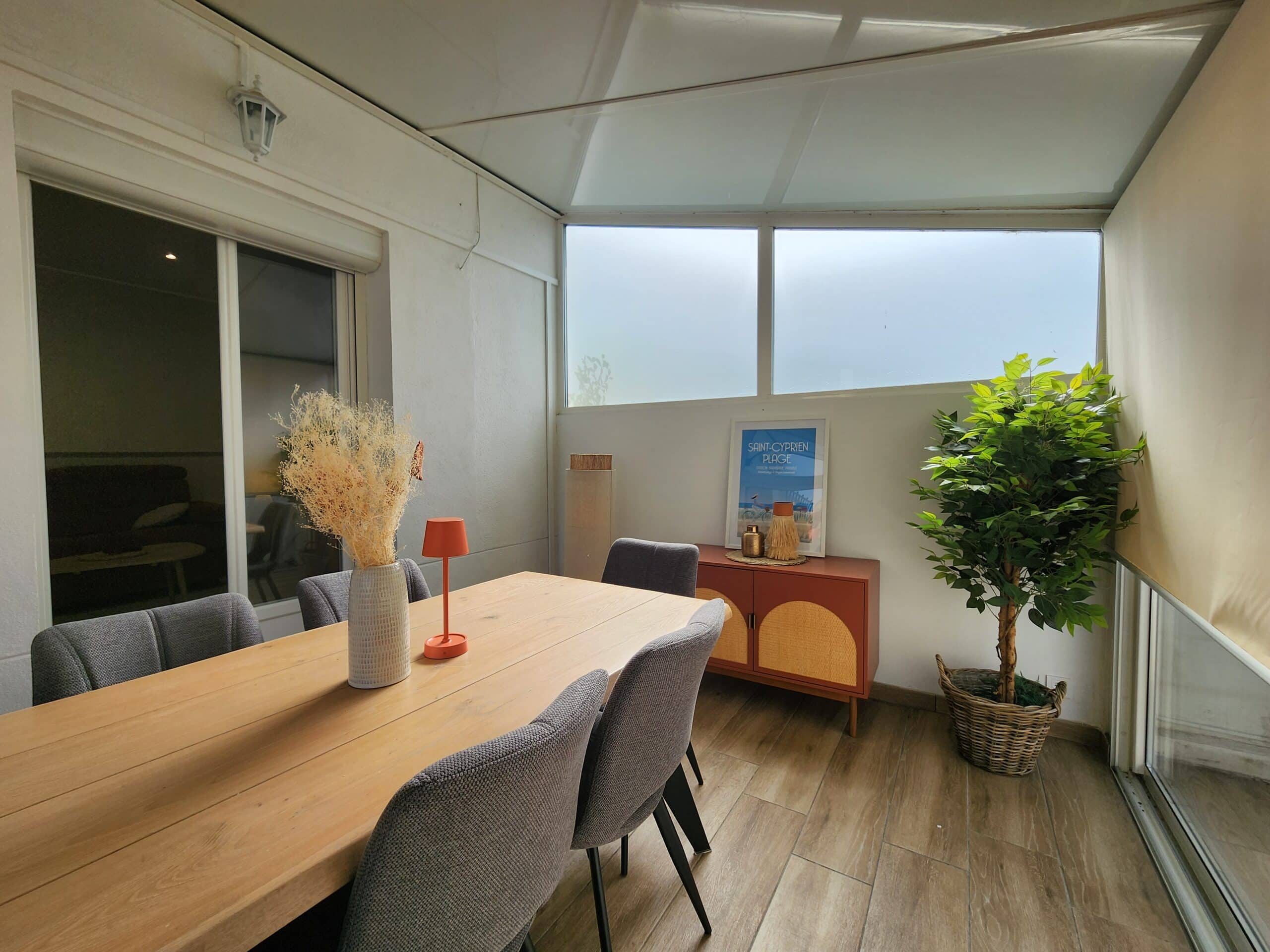 Salle à manger lumineuse : table bois, chaises grises, buffet terracotta et affiche "Saint-Cyprien Plage".