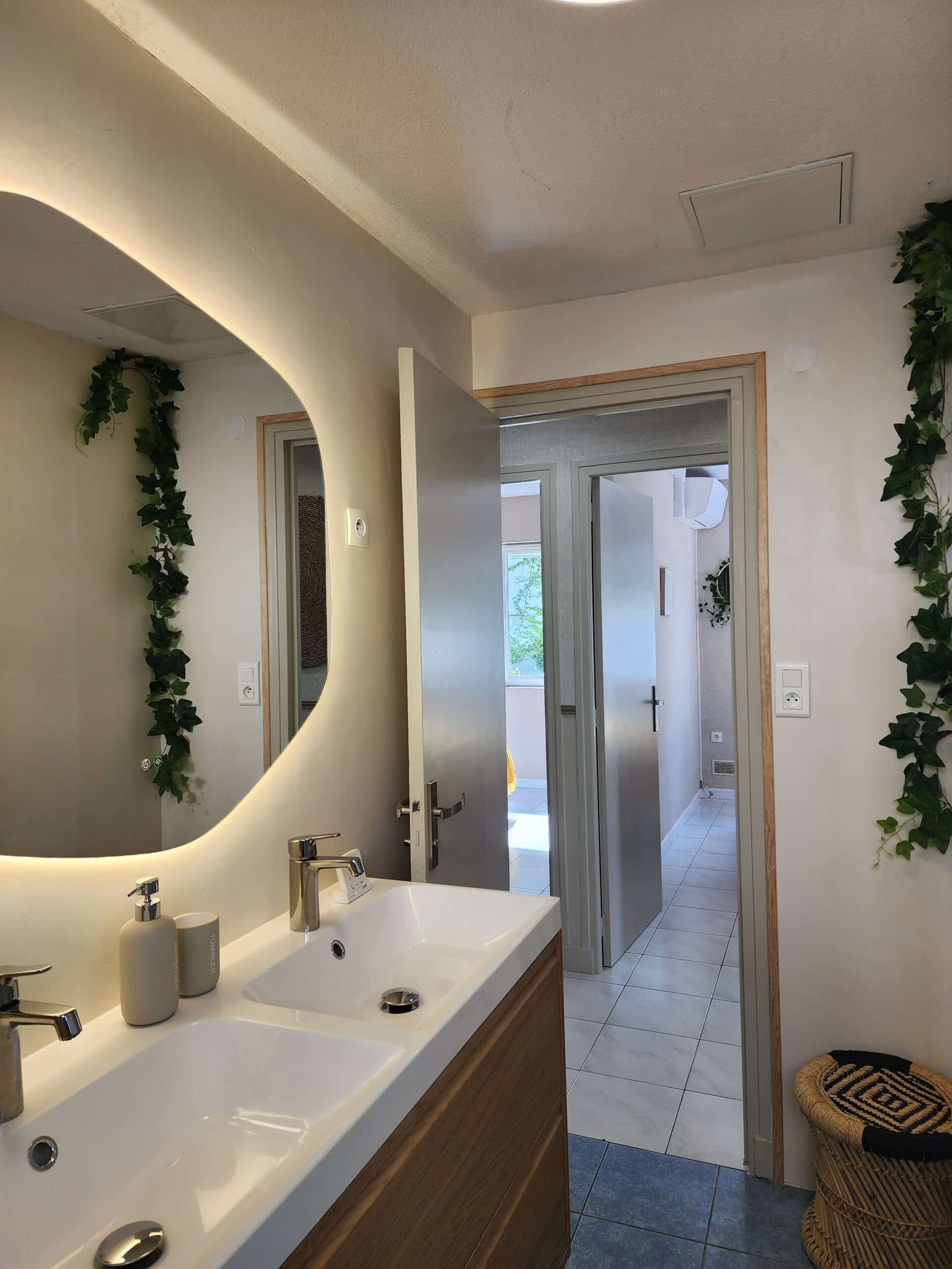 Salle de bain moderne : double vasque blanche, miroir ovale rétroéclairé et lierre décoratif mural.