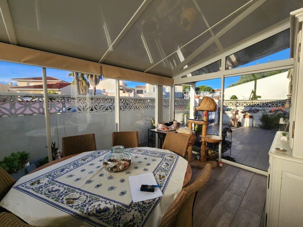 Véranda lumineuse avec table ronde, chaises en osier et arbre à chat face à une terrasse ensoleillée.