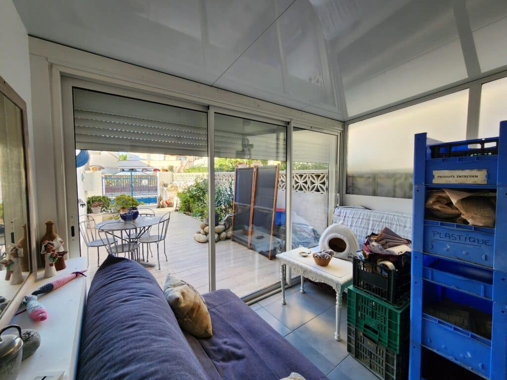 Salon de véranda avec canapé, vue sur patio. Caisses 'Plastique' et 'Produits entretien' incluses.