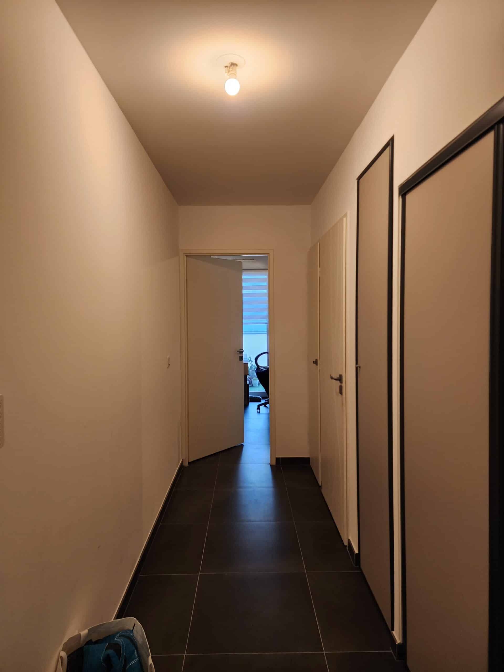 Couloir moderne aux murs blancs, carrelage noir et portes de placard, éclairé par un plafonnier central.