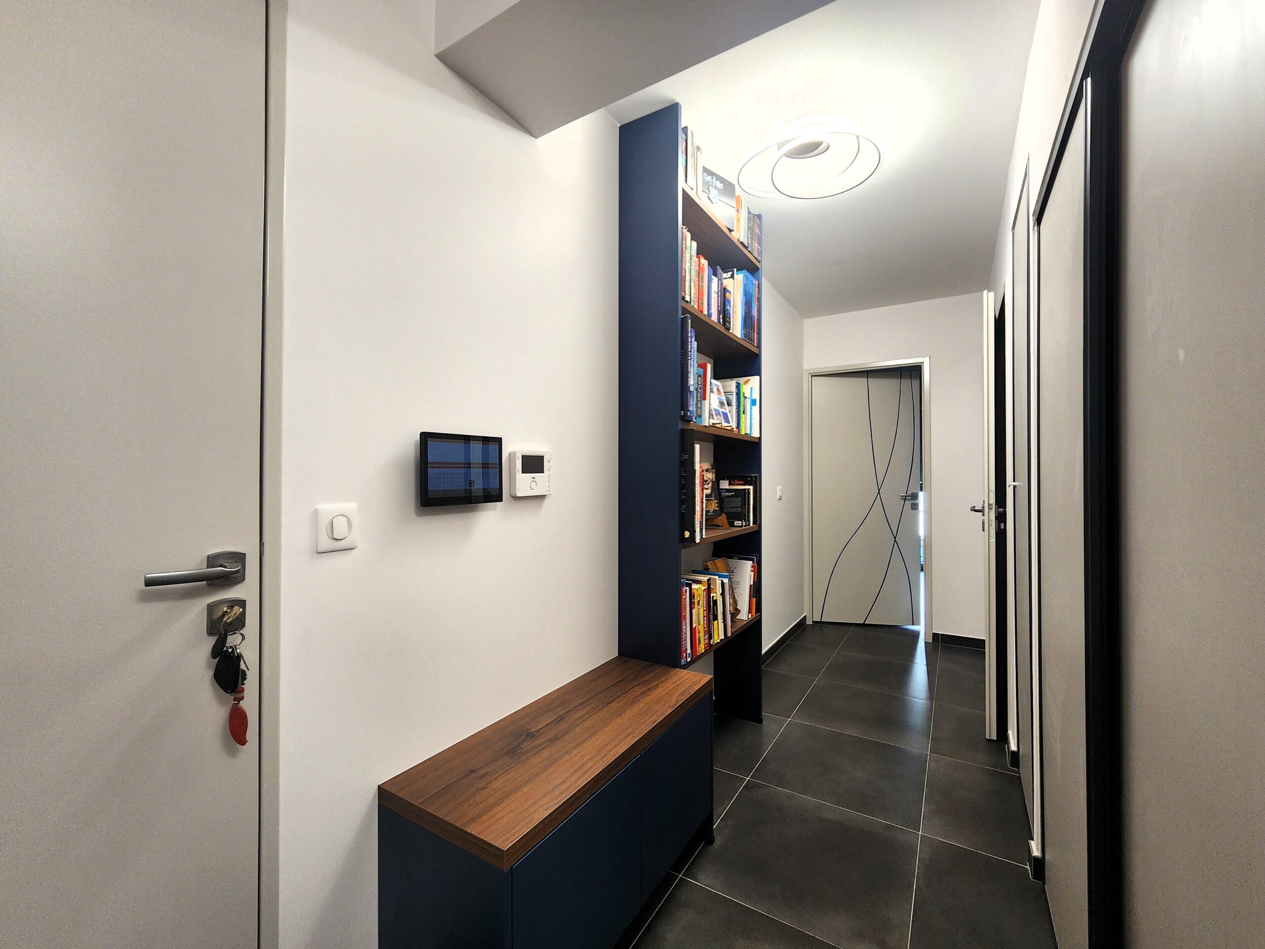 Entrée moderne avec meuble bleu et bois, tablette domotique et carrelage gris dans un couloir au design épuré.
