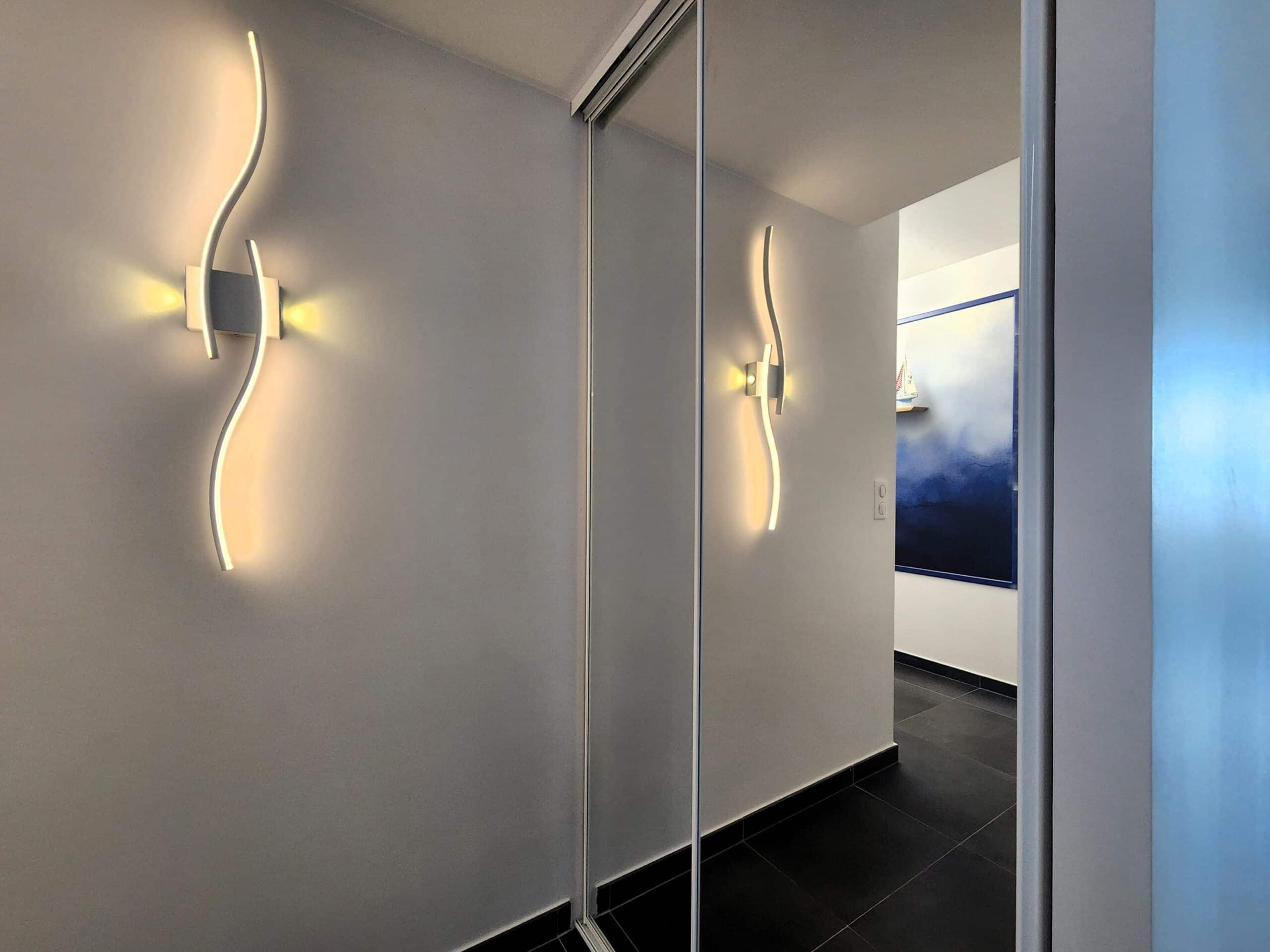Applique murale LED design sinueuse sur mur blanc se reflétant dans un miroir de placard de couloir moderne.